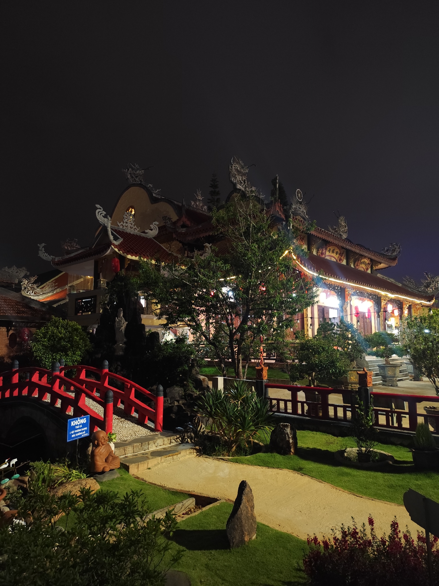 This image presents a serene night view of a beautifully illuminated temple complex, likely located in Lâm Viên - Đà Lạt, Vietnam. The scene is devoid of people, contributing to a peaceful and contemplative atmosphere.
The primary subject is a multi-tiered traditional temple building, showcasing intricate East Asian architecture. Its reddish-brown tiled roofs are adorned with ornate carvings and sculptures, possibly of dragons or mythical beasts, which are highlighted by warm lights. The facade of the temple is brightly lit, revealing detailed wooden doors and windows, and is further enhanced by decorative string lights and traditional red lanterns hanging from the eaves.
In the foreground, a meticulously maintained garden features vibrant green grass and beige pathways. A prominent red arched bridge with matching railings curves gracefully over a path, leading towards the main temple structure. Near the bridge, a small, brown terracotta statue of a seated monk or Buddha figure is placed. Adjacent to this statue, a blue sign displays Vietnamese text: "KHÔNG BẮT CÁ (VIỆT HÔNG SÁNH) VỚI XUỐNG HỒ," which translates to "DO NOT CATCH FISH (or touch the fish) OR GO DOWN TO THE POND," indicating rules for visitors.
The garden also contains various lush trees and bushes, some subtly adorned with string lights, and several large, natural rocks positioned throughout the landscape. Amidst the foliage closer to the temple, a white statue, possibly depicting a deity such as Guan Yin, is visible. The dark night sky provides a striking contrast to the warmly lit temple and garden, emphasizing the intricate details and creating a tranquil, almost magical ambiance. The setting suggests a well-cared-for religious site, possibly open to visitors for evening contemplation or sightseeing.