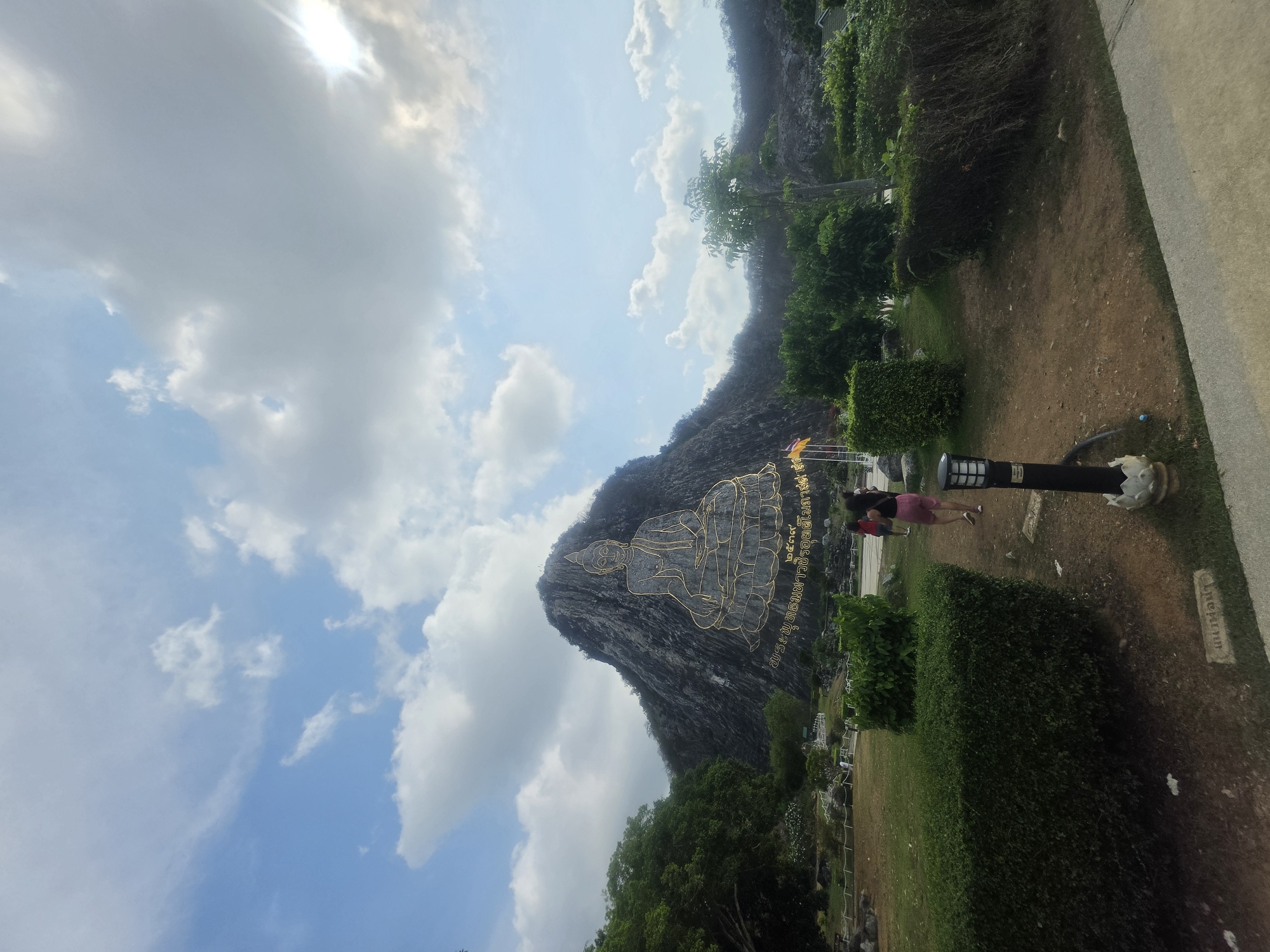 The image captures the majestic Khao Chi Chan Buddha Mountain (Buddha Mountain) in Bang Sare, Thailand, under a bright, partially cloudy sky. The main subject is a colossal golden outline of a seated Buddha, laser-carved onto the face of a greyish-brown limestone mountain. The Buddha image depicts the Maravijaya mudra, symbolizing victory over temptation.
To the right of the Buddha carving, Thai script is etched into the mountain, reading "พระพุทธมหาวชิรอุตตโมภาสศาสดา" (Phra Phuttha Maha Wachira Uttamophat Satsada), which is the official name of the Buddha, followed by "๒๕๓๙" (2539 BE), indicating the year 1996 CE.
In the foreground and midground, lush green vegetation including trees and manicured bushes frames the scene. A dirt path and a paved walkway are visible. Two people, an adult in a red top and pink shorts, and a smaller figure, are standing in the mid-right, appearing to observe the landmark. A black lamppost with a white decorative base stands nearby.
The bright daylight is evident from the clear blue sky with scattered white clouds, and a noticeable sun flare in the upper left portion of the sky. The overall scene suggests a visit to a popular and significant cultural and religious site on a pleasant day.
