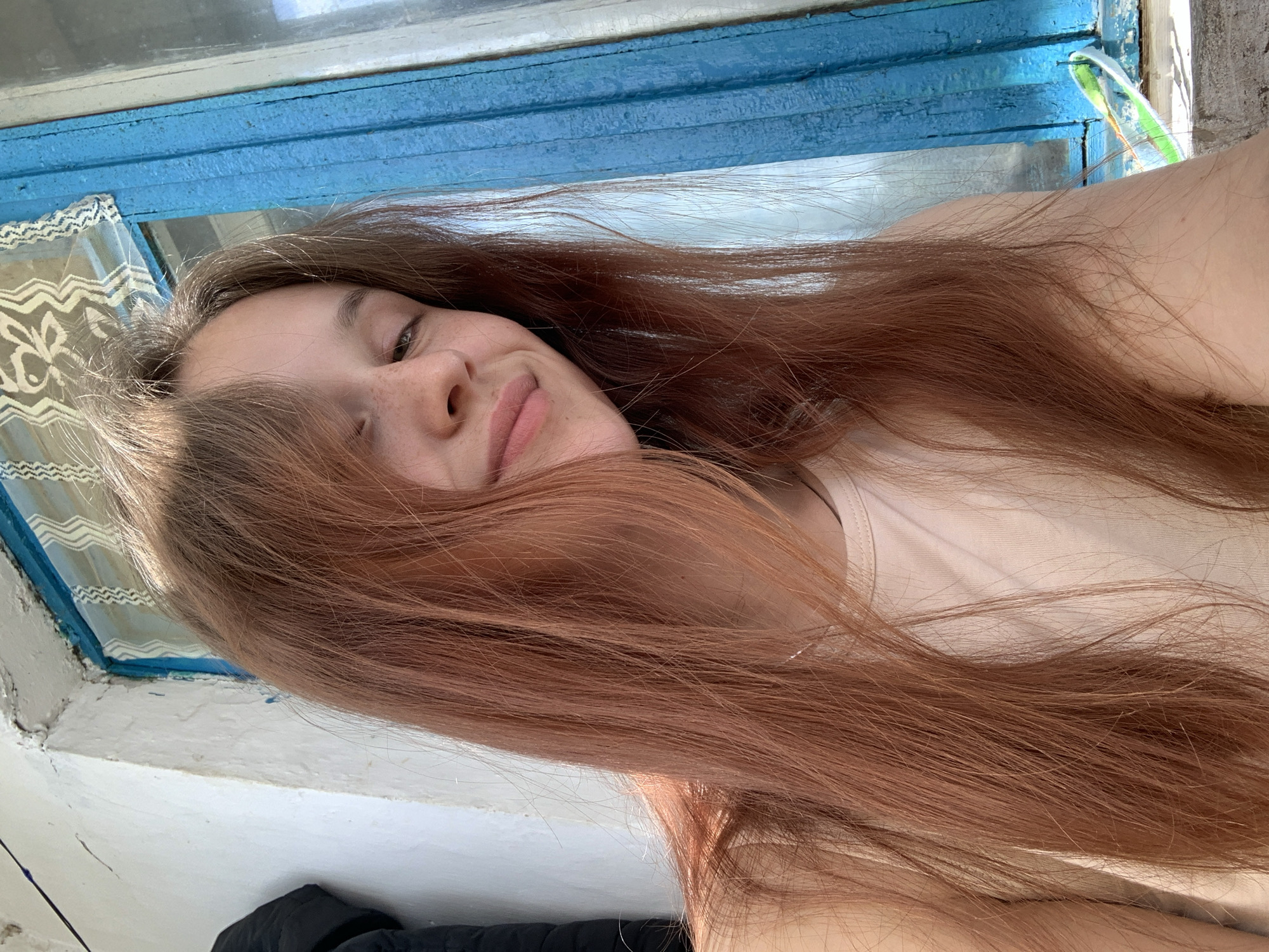 A young woman with long reddish-brown hair lies near a window. The window frame is painted blue and the window has a lace curtain. A black object is visible in the lower-left corner. The woman is smiling slightly. The scene appears to be indoors, near a window, but the precise location in the city of Фоки, Russia, cannot be determined from the image.