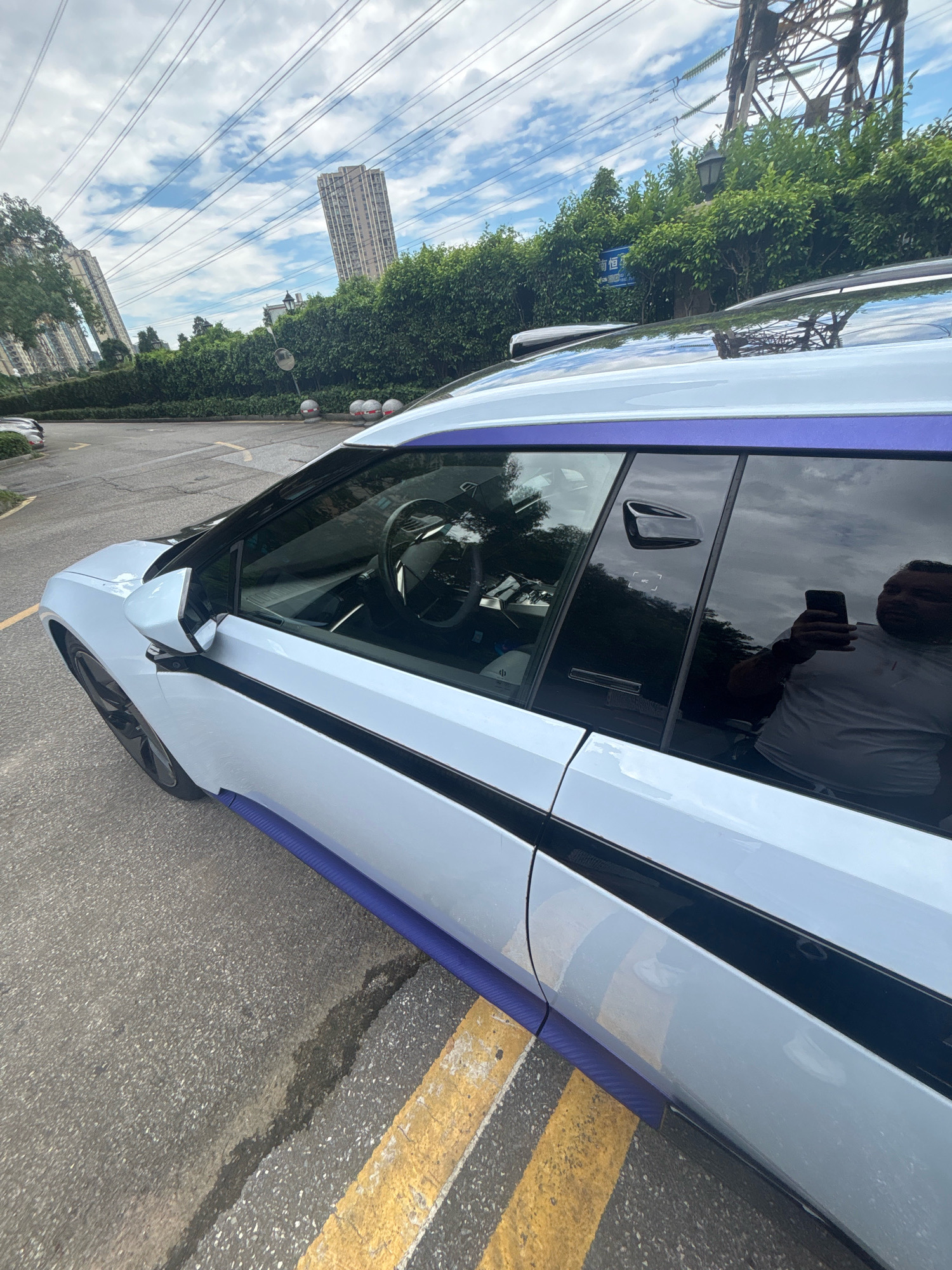 An outdoor, eye-level, medium shot captures the passenger side of a white electric vehicle parked on a paved road with yellow and white painted lane markings. The car's doors are closed, and its tinted windows reflect the cloudy sky and surrounding trees. A person is visible through the rear passenger window, holding a smartphone and appearing to take a photograph. In the background, several tall, modern buildings rise against a partly cloudy sky, with overhead power lines and a large industrial structure dominating the upper portion of the frame. The environment suggests an urban setting in the Nanhai District of China.