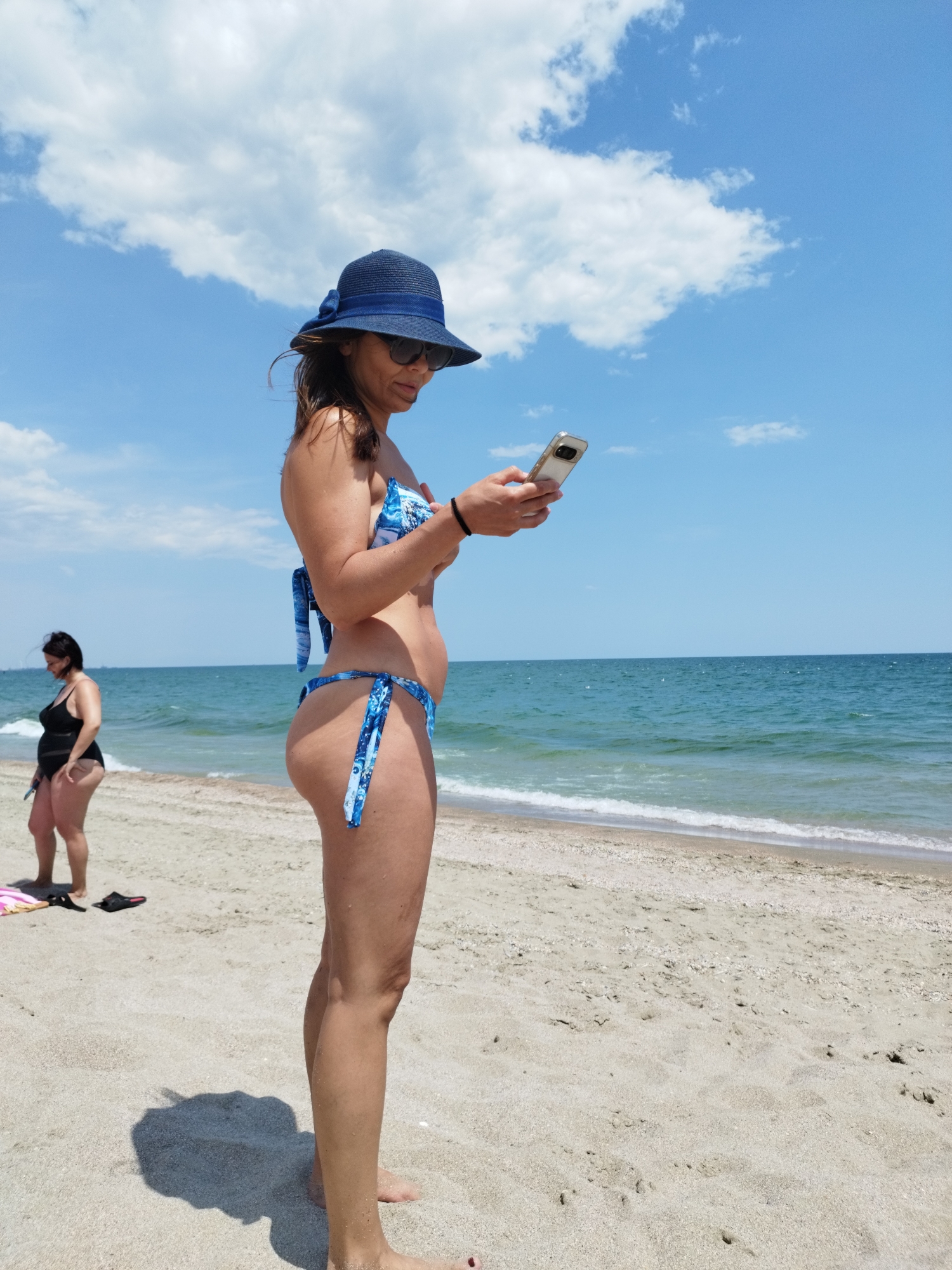 The image shows a beach scene on a sunny day. In the foreground, a woman in a blue and white patterned bikini and a blue hat is standing on the sand, looking at her phone. She is wearing sunglasses. To the left and further back, another woman in a black swimsuit is standing on the sand. The sea is visible in the background, with waves gently rolling onto the shore. The sand is light in color and appears dry. The sky is blue with scattered white clouds. Based on the context of Constanța, Romania, this scene is likely depicting a day at one of its beaches.