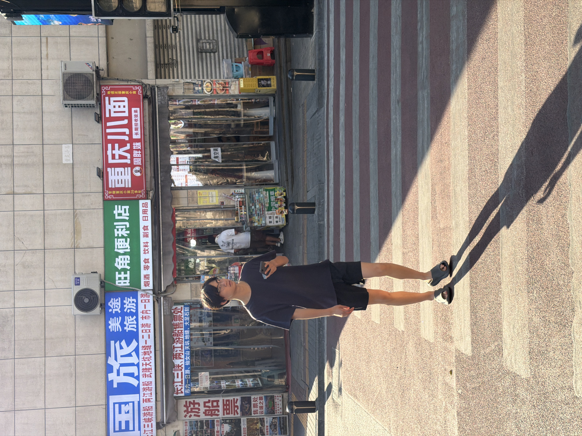 A young East Asian male with short dark hair and glasses is walking across a crosswalk in the Yuzhong District of Chongqing, China. He is wearing a dark gray t-shirt and black shorts, and dark gray slides. He is holding a mobile phone in his right hand. The sun is casting a long shadow of him across the crosswalk, indicating it is late afternoon or early morning. Behind him, a row of storefronts is visible, including signs for "Chongqing Xiaomian" (a type of noodle dish), a travel agency, and a convenience store. The street appears to be a busy urban area.