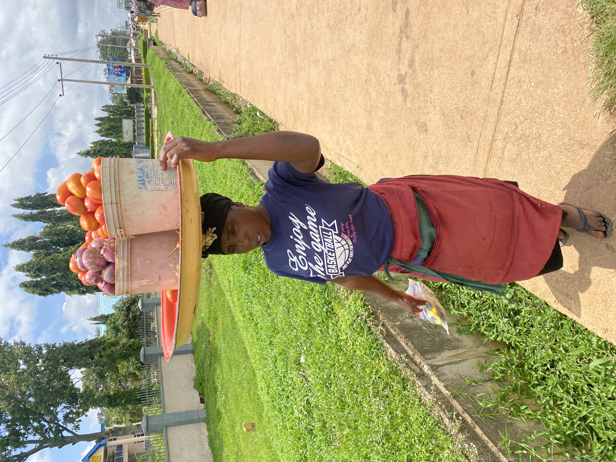 In Bukuru, Nigeria, a woman walks along a paved path, carrying a large red and yellow tray balanced on her head. On the tray are two buckets filled with produce. The bucket on the left contains red onions, and the bucket on the right is filled with tomatoes. The woman is wearing a dark blue t-shirt with white lettering that reads "Enjoy the game BASKETBALL," a red skirt, and dark sandals. She is holding a clear plastic bag with yellow items in her left hand. The path is lined with green grass and a concrete drainage ditch. In the background, there are trees, buildings, and utility poles, with a cloudy sky overhead.