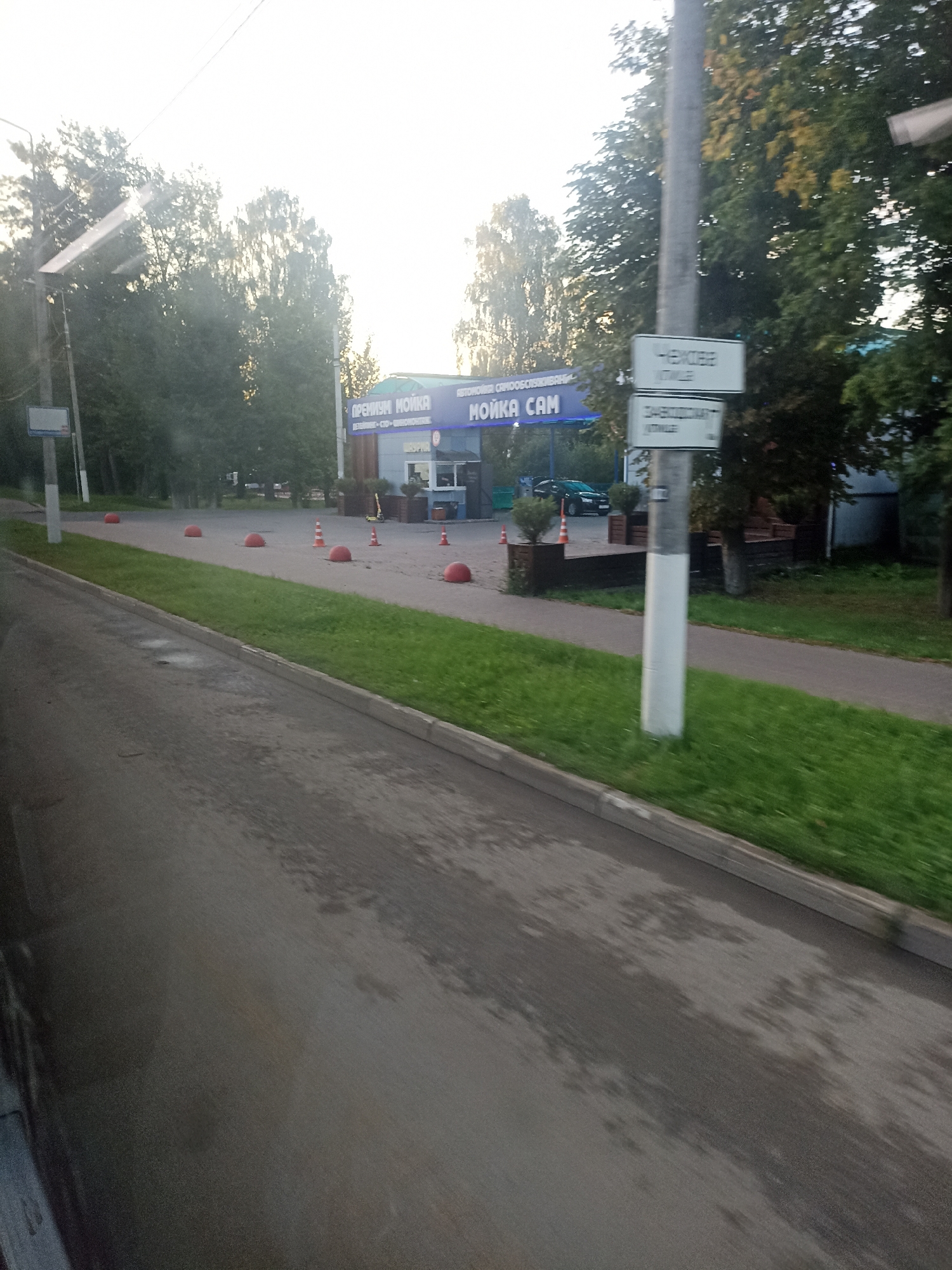 A car wash sign reads "PREMIUM MOYKA" in blue text, next to "MOYKA SAM" in white text. A light pole stands nearby with two signs attached. The top sign reads "CHEKHOV" and the bottom reads "ZAKHODIMYY" which translates to "ENTRANCE". The car wash is located on a street in Chekhov, Russia. The street is paved, with a grassy area on the side, and several red bollards are placed along the curb. Trees line the background, and the sky is bright with sunlight.