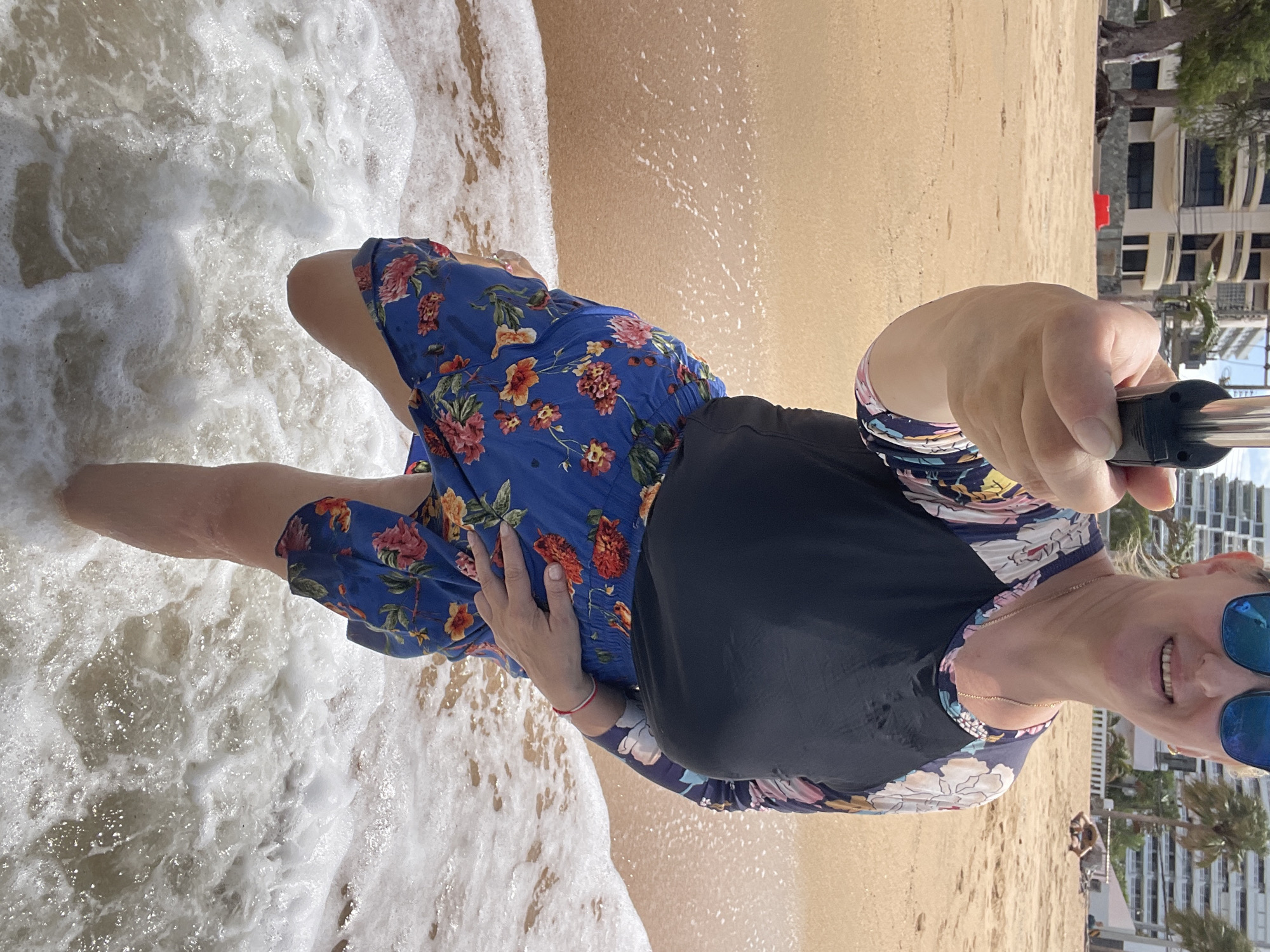 A woman stands on a sandy beach in Pattaya City, Thailand, with gentle waves washing over her feet. She is holding a selfie stick, taking a photo of herself. She wears a long-sleeved rash guard with a floral pattern on the sleeves and a solid navy blue front, paired with blue shorts adorned with a colorful floral print. The backdrop features a sandy beach, the turquoise ocean, and several buildings in the distance, including a multi-story hotel. Palm trees are visible, adding to the tropical ambiance. The sun appears to be shining, casting a bright light on the scene.