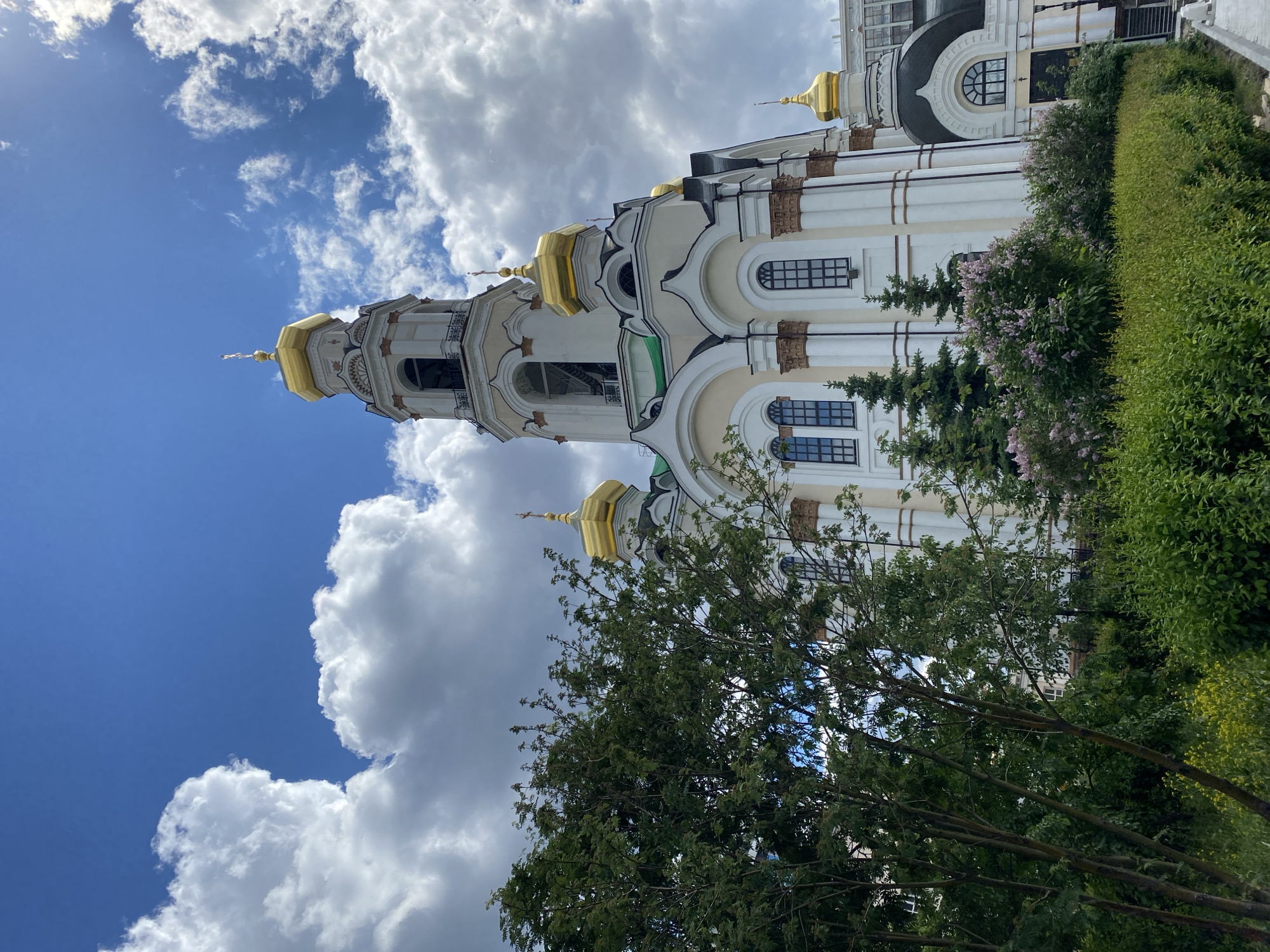 The image depicts a tall, ornate building with golden domes, likely a church or cathedral, set against a partly cloudy sky. The architecture features light-colored walls, arched windows, and decorative elements. Green trees and shrubs surround the building, partially obscuring the lower sections. The scene is likely located in the city of Yekaterinburg, Russia.