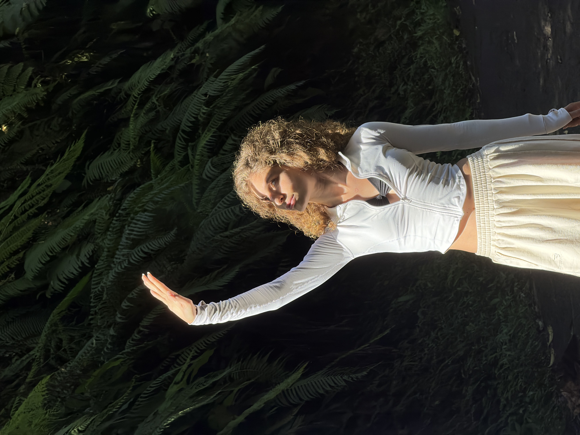 A young woman with curly blonde hair stands in front of a dense green wall of ferns. She is wearing a white zip-up long-sleeve shirt, a black bralette, and loose cream-colored pants. Her right arm is raised, with her hand shielding her face from the sun. The sunlight illuminates her face and arm, creating a strong contrast with the shadowed background. The image appears to be taken outdoors, possibly in a tropical or humid environment, such as the location in Penebel, Indonesia, known for its lush natural landscapes and rice terraces.