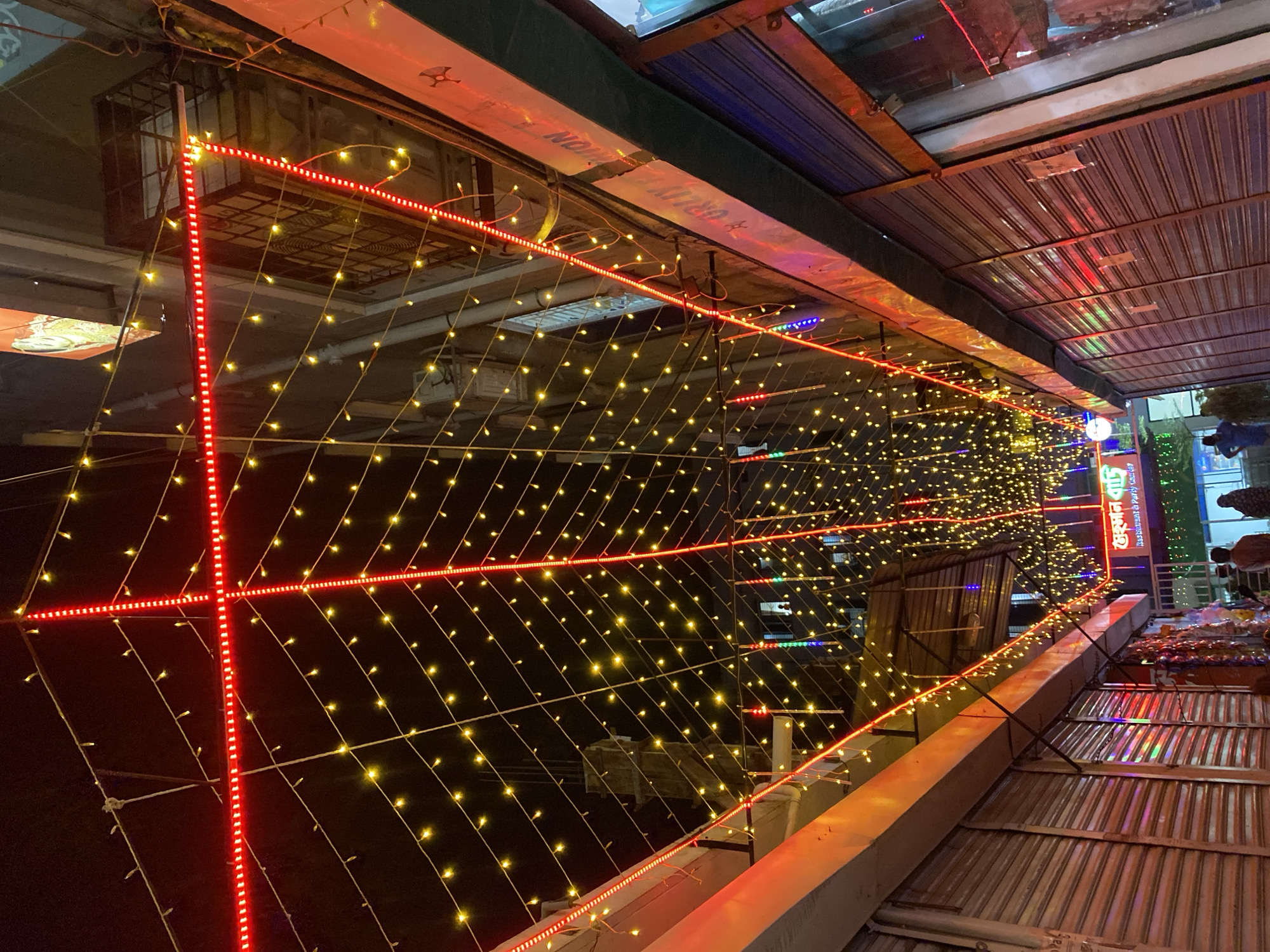 The image captures an illuminated outdoor market scene in Dhaka, Bangladesh at night. A dense grid of small, warm-toned lights is the dominant feature, hanging as a decorative canopy. This grid is accented by linear runs of bright red LED lights, forming geometric shapes within the larger field.
Above, a metal corrugated roof extends over the market area, reflecting some of the light. Below, a concrete structure or wall forms a base for the lighting display.
In the background, signs in Bengali script are visible, and glimpses of shopfronts can be seen. The overall impression is of a busy commercial area lit for evening trade. There are no people visible in the image, but goods displayed at the periphery indicate active commerce.