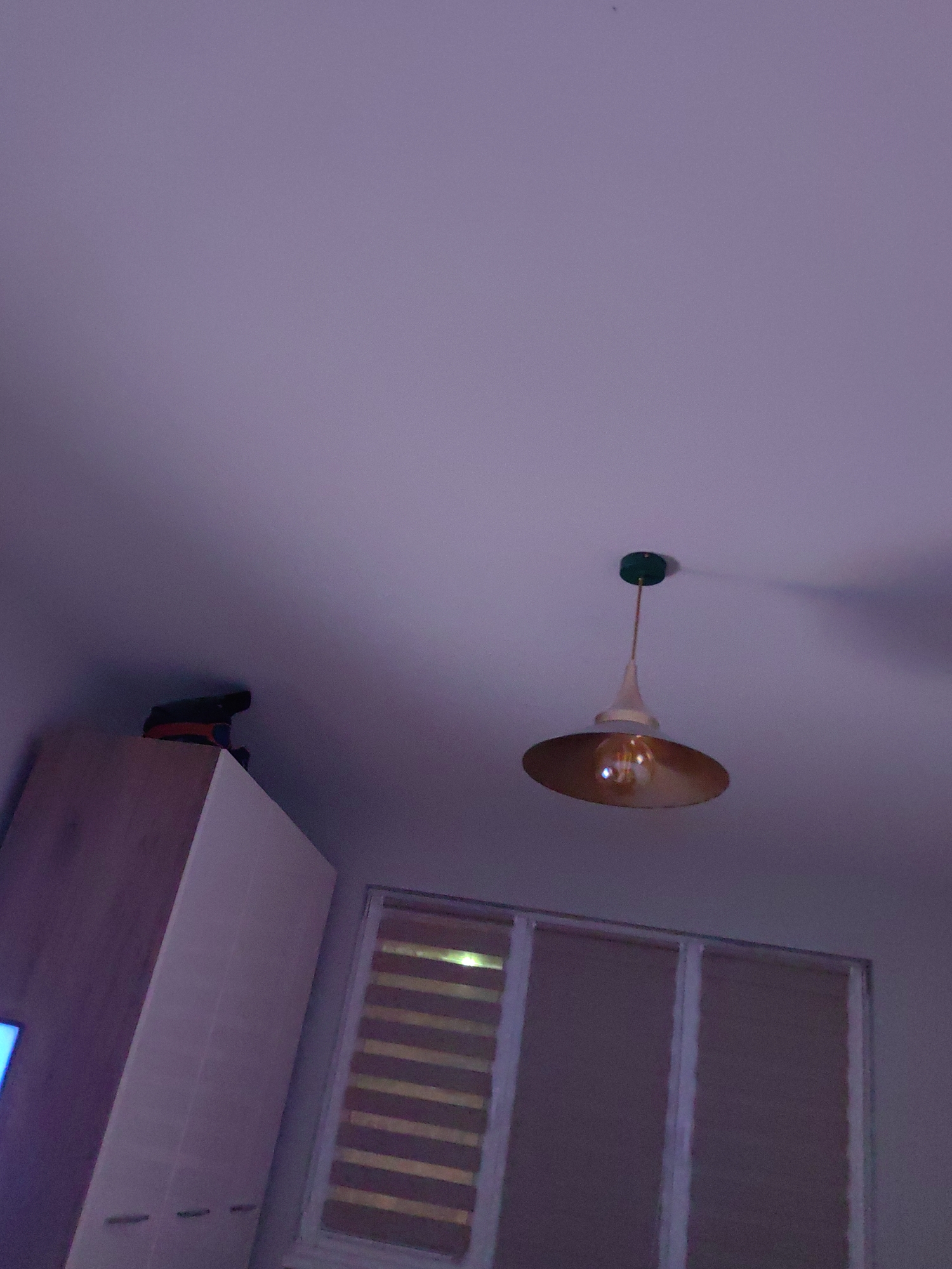 The image presents an indoor scene, likely a bedroom. Dominating the view is a ceiling-mounted light fixture with a golden-colored lampshade, a white conical component, and a green mounting base. Below the light, a window with partially drawn blinds is visible. To the left of the window, a white wardrobe extends vertically. On top of the wardrobe, a small dark object is perched. The overall color cast of the image is purplish, likely due to the lighting conditions. The location is unknown, and there is no specific information to indicate the image was taken in Sofia, Bulgaria. The scene appears static, with no immediate actions or interactions evident.
