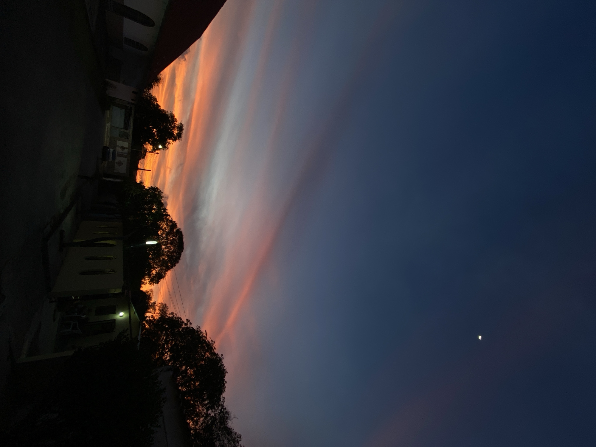 The image captures an evening scene at dusk in Juan Griego, Venezuela. The sky displays a gradient, transitioning from a dark blue overhead to vivid orange, red, and pink cloud formations near the horizon. A crescent moon is visible in the upper right quadrant. The foreground depicts an unlit paved street. On the far left, a structure with a reddish-brown gabled roof and light-colored walls featuring dark, arched window apertures is partially visible. Along the right side of the street, multiple buildings are present. One building exhibits a light-toned facade, an illuminated exterior wall lamp, and two white plastic chairs positioned near its entrance. A light-colored sedan is parked further down the street on the right. Dense, dark tree canopies are prominent across the midground and right, silhouetted against the sunset. A utility pole with power lines is also visible above the structures. No human figures are present, and no active interactions are depicted.