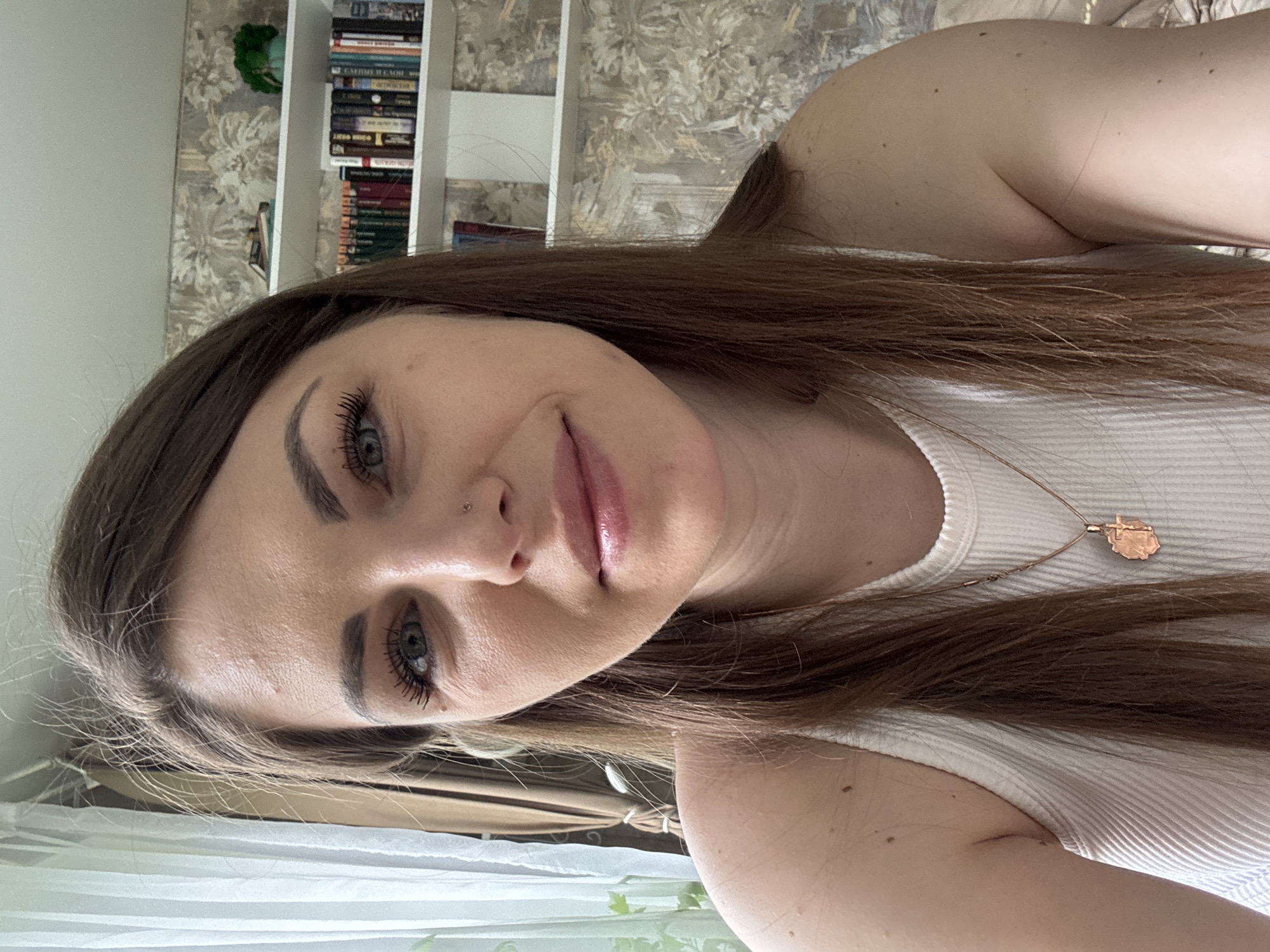 A close-up selfie of a young woman with light skin and brown hair is presented. She has a nose piercing and wears noticeable mascara. A thin gold necklace adorns her neck. She is wearing a white ribbed tank top. In the background, a bookshelf filled with books is visible. The wall has a floral pattern. The image may not depict Раздолье, Russia, as there are no location-specific details.