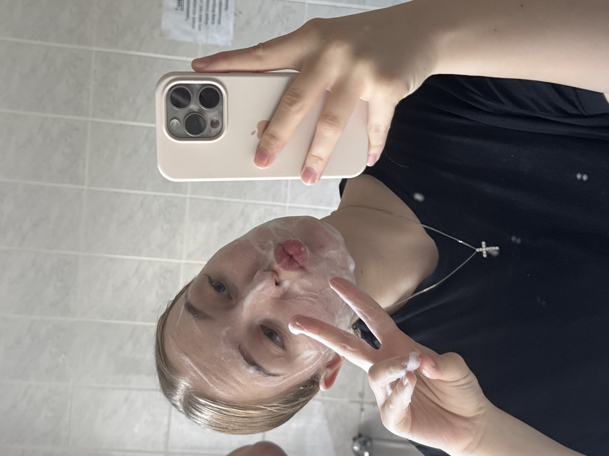 A young woman is captured in a mirror selfie, taken in what appears to be a bathroom. Her face is covered in a white, foamy substance, likely a facial cleanser or mask. She is looking directly at her reflection, making a "duck face" with her lips and holding up a peace sign with her left hand, which also has some of the white substance on it. She has light-colored hair, pulled back, and is wearing a black t-shirt with a silver cross necklace.
The selfie is being taken with a light pink or beige iPhone, visible in her right hand, featuring a prominent three-lens camera system on the back. The background consists of light-colored, possibly white or very light gray, square tiles, characteristic of a bathroom. A faint reflection of what looks like a sink or faucet is visible at the bottom right corner of the image. In the top left corner, reflected in the mirror, is a piece of paper with unreadable text. The location of the scene is Perm, Russia. The overall mood is casual and playful.