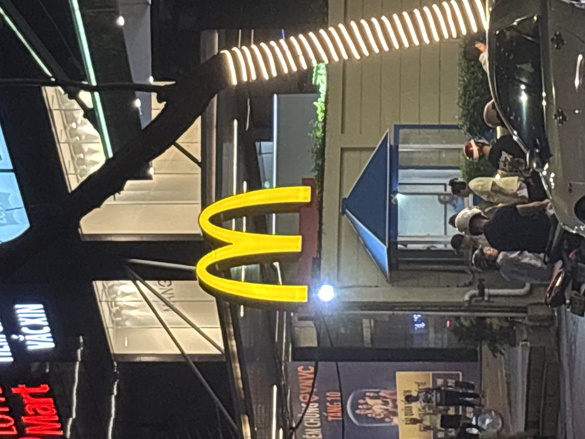 The image captures an outdoor scene at night, likely in Nha Trang Ward, Vietnam, featuring a prominent McDonald's restaurant. The iconic yellow M arches of the McDonald's sign are illuminated and clearly visible. In the foreground, several people are gathered, some on motorcycles, suggesting a bustling urban environment. A car is parked nearby. The background shows the facade of the McDonald's building, with windows and architectural details. Signs in Vietnamese are visible on the building, indicating local business names and possibly directions. The overall atmosphere is that of a lively commercial area at night, illuminated by artificial lighting from signs and buildings.