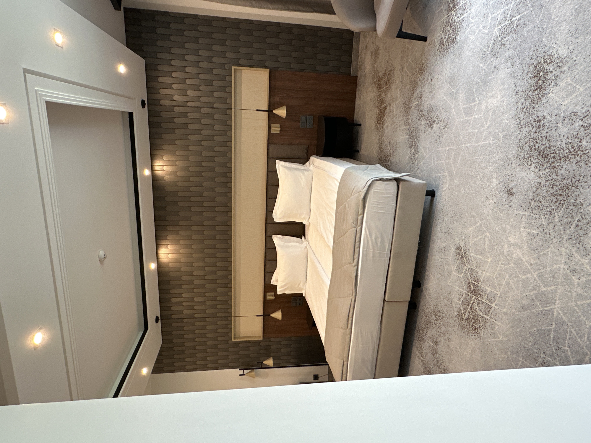 This is an interior shot of a bedroom. The main focal point is a neatly made bed with white linens, positioned centrally in the frame. The headboard is a combination of light-colored rectangular panels and a darker, wood-like surface with integrated lighting fixtures. A gray and white patterned carpet covers the floor. Part of a gray armchair is visible in the upper right corner. The left side of the image features a white wall with a doorway, illuminated by embedded lights. The room's design elements, including the headboard and wall coverings, suggest a modern aesthetic. The exact location within the city of Kazanlak, Bulgaria, cannot be determined from this image alone.