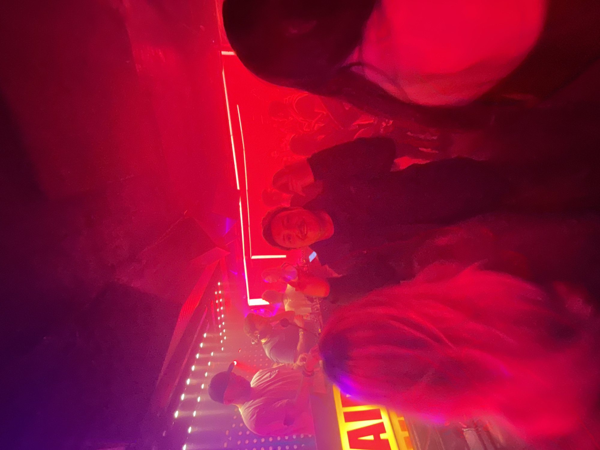 The image shows a group of people illuminated by red and violet light, seemingly in a nightclub or bar. The location is Mexico City, Mexico. A man in a black jacket smiles at the camera. Several other individuals are partially visible, some holding drinks. The environment has decorative lighting. A yellow sign with the letters "AI" is visible in the foreground.