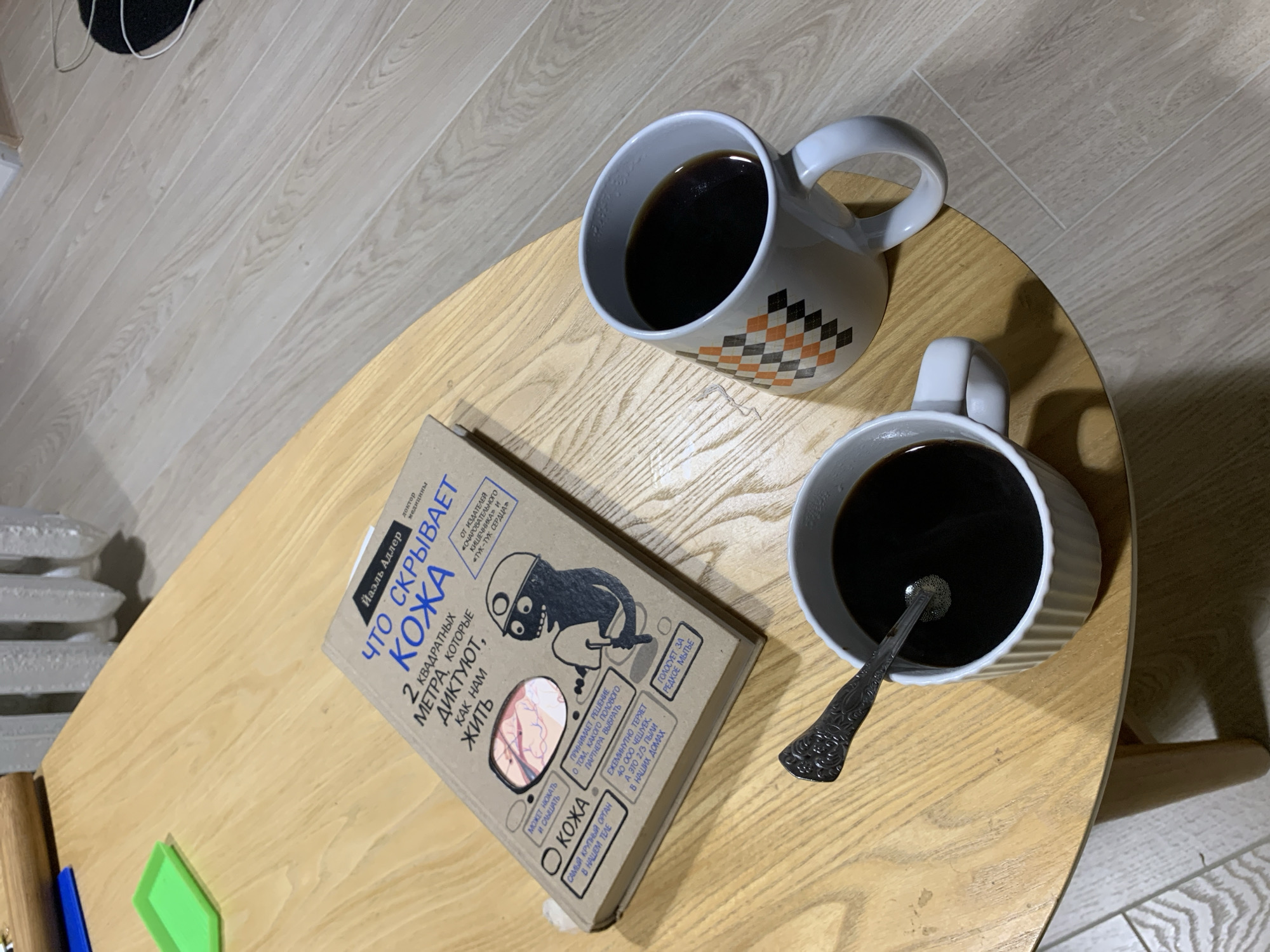 The image shows a light wood-colored, oval coffee table with two mugs of dark liquid, presumably coffee, and a book. One mug is white with a geometric pattern of orange, black, and white squares. The other mug is white and fluted, with a silver spoon inside. The book is titled "Что скрывает кожа" (What the Skin Hides) by Илль Адлер. The book is lying open, revealing some text and illustrations. A green object, possibly a small plastic container, is visible on the table to the left of the book. The background shows a light-colored wooden floor and a white radiator. The scene appears to be indoors, possibly in a residential setting in the city of Kapyĺ, Belarus. There are no people visible in the image.
