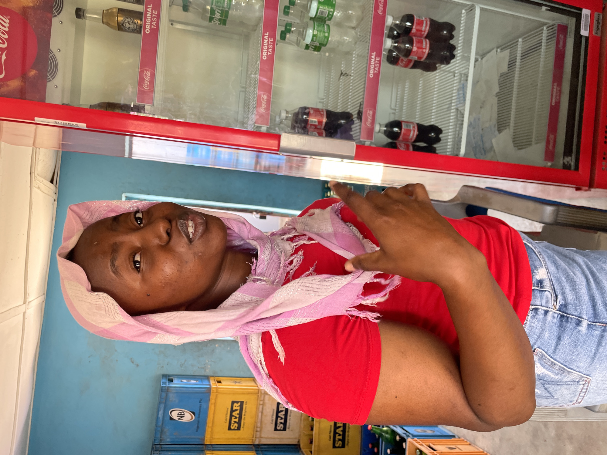 A medium shot captures a woman in a red shirt and denim bottoms, adorned with a pink patterned scarf. She stands indoors, next to a refrigeration unit stocked with beverages, including Coca-Cola and Sprite. Stacks of blue and yellow bottle crates labeled "STAR" and "NB" are visible. The environment suggests a commercial establishment, likely a shop or kiosk. The location is suggested by the presence of Nigerian Breweries crates, but the city of Jos is not visually confirmable.