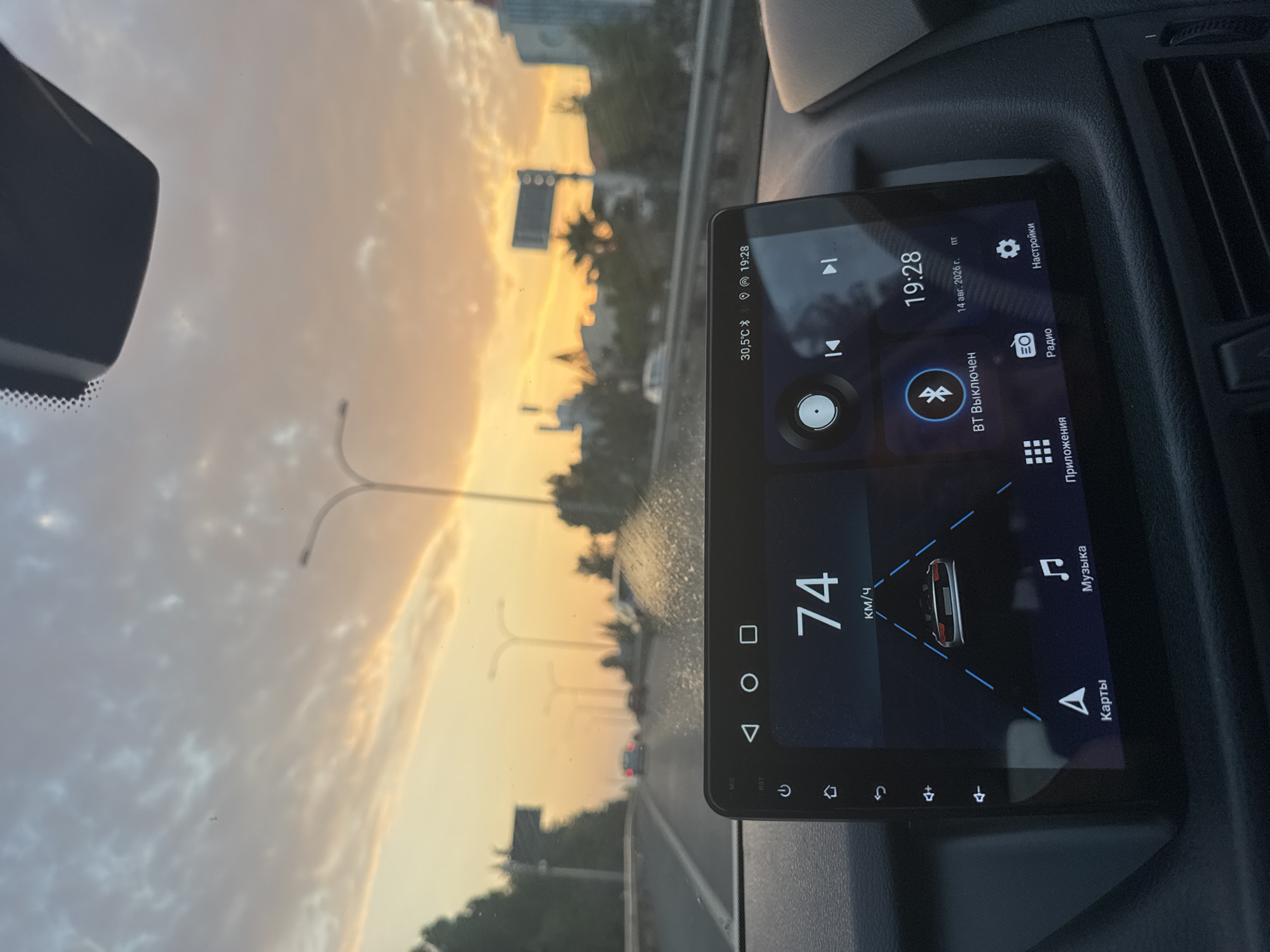 An interior shot from a vehicle's perspective shows a dashboard screen displaying navigation and media information against a backdrop of a sunset driving on a highway. The screen shows a speed of 74 km/h, a visual representation of a car on the road, and icons for "Карты" (Maps), "Музыка" (Music), "Приложения" (Applications), and "Радио" (Radio). The top bar of the screen shows the temperature as 30.5°C, a Bluetooth icon indicating it is disconnected ("BT Выключен"), and the time 19:28 along with the date "14 авг. 2026 г." (August 14, 2026). The sky is filled with orange and pink clouds, characteristic of a sunset. The road ahead is visible with streetlights and other vehicles in the distance. The location is identified as Agios Athanasios Municipality, Cyprus.