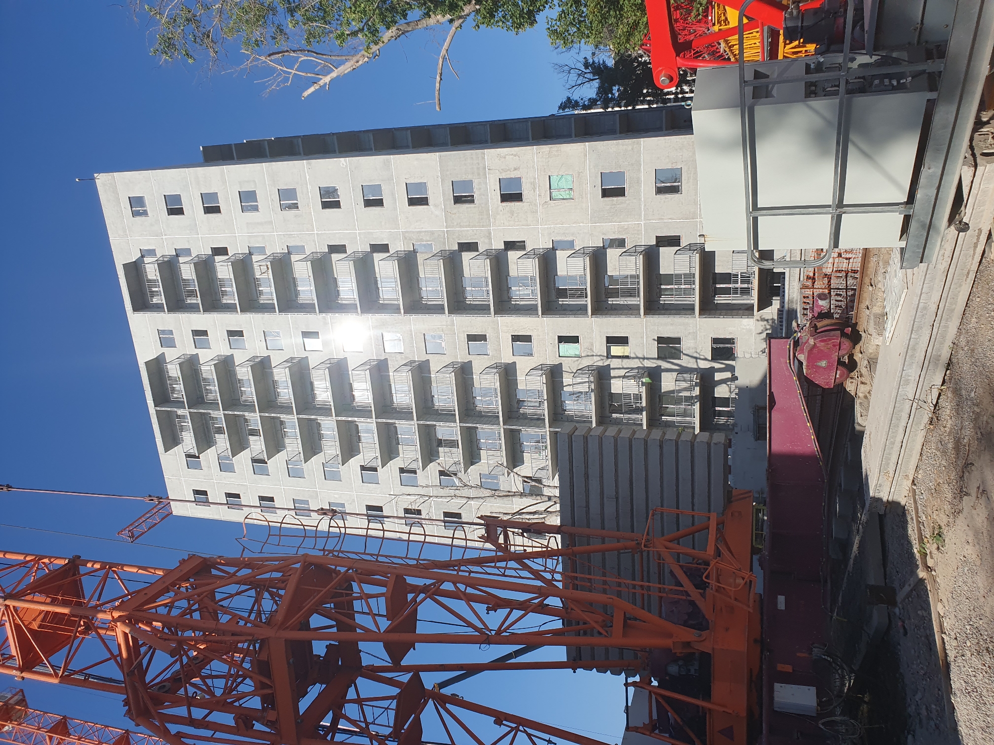 An unfinished tall residential building under construction is viewed from a low angle against a clear blue sky. An orange crane is prominently positioned on the left, its lattice structure dominating the foreground. Large precast concrete slabs are stacked in front of the building's base. The building itself has a concrete facade with numerous balconies, each with black metal railings. Many windows are visible, some are open, and a few have green protective coverings. Construction equipment, including a red crane arm, is visible in the lower right corner and in the background. The ground in the foreground is covered with gravel and debris. This scene is likely from a construction site in Tashkent, Uzbekistan, given the presence of construction on such a scale in a growing urban environment.
