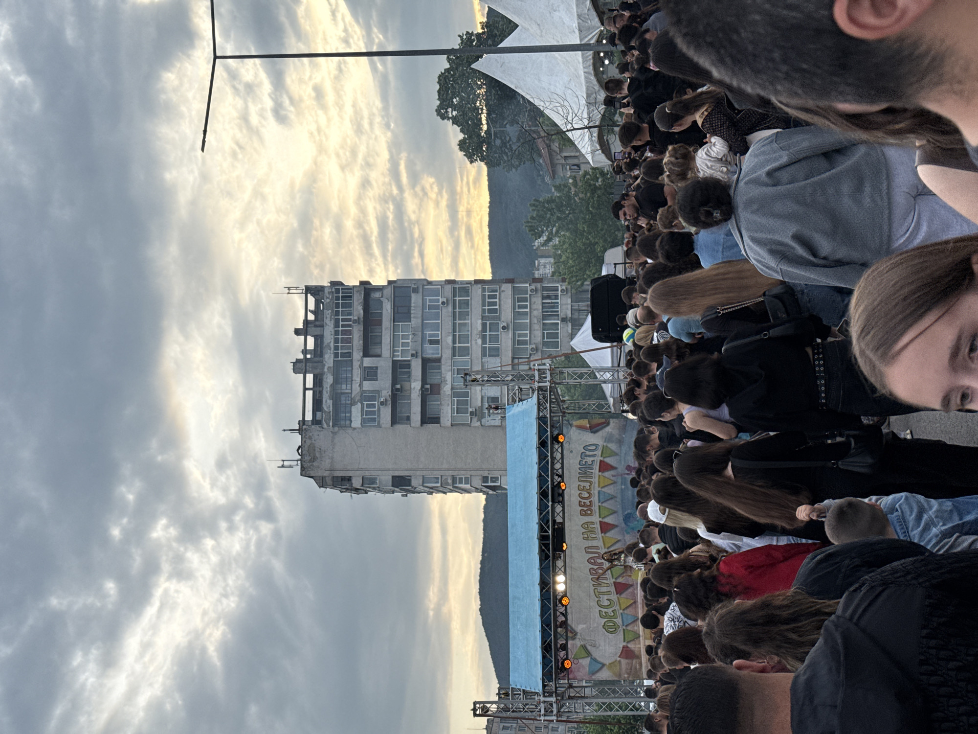 This image captures an outdoor festival or concert in Shumen, Bulgaria, during the late afternoon or early evening. A large crowd of people occupies the foreground and midground, mostly viewed from behind, as they face a performance stage. Many attendees have dark hair and are casually dressed.
In the midground, a large stage is set up with a blue canopy roof and a colorful banner forming its backdrop. The banner displays Cyrillic text: "ФЕСТИВАЛ НА ВЕСЕЛЕТО," which translates to "FESTIVAL OF MERRIMENT" or "FESTIVAL OF FUN." Speakers and various stage lighting elements are visible on the metal scaffolding supporting the structure.
The background features a prominent, tall, grey multi-story apartment building, indicating an urban setting. Behind this building, distant hills or mountains rise, covered with trees. The sky is overcast with a mix of dark and brighter clouds. The low-angle sunlight, likely from the setting sun, casts a warm, soft glow across the horizon and illuminates parts of the cloud cover. A tall streetlight pole stands on the left side of the frame. The scene conveys a lively public event taking place under a beautiful, dramatic sky.