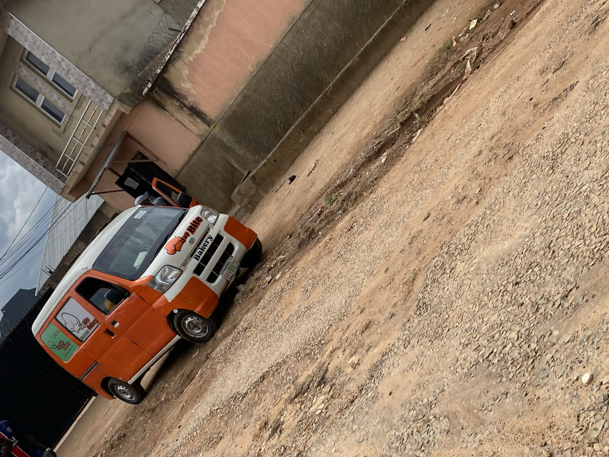 An orange and white van, labeled "Jay Bite Bakery," is parked on a dirt road in Rafin Par, Nigeria. The van has a baking graphic and the words "Bakery" on the front. The van's license plate reads "BKK-99110." A person wearing a white cap and blue garment is partially visible inside the van, near the open driver's side door. A building with a balcony and grey tiled accents is situated to the right of the van, and a barbed wire fence runs along the front of the building. The road is unpaved and covered in gravel and dirt, with some scattered debris. The sky is overcast.