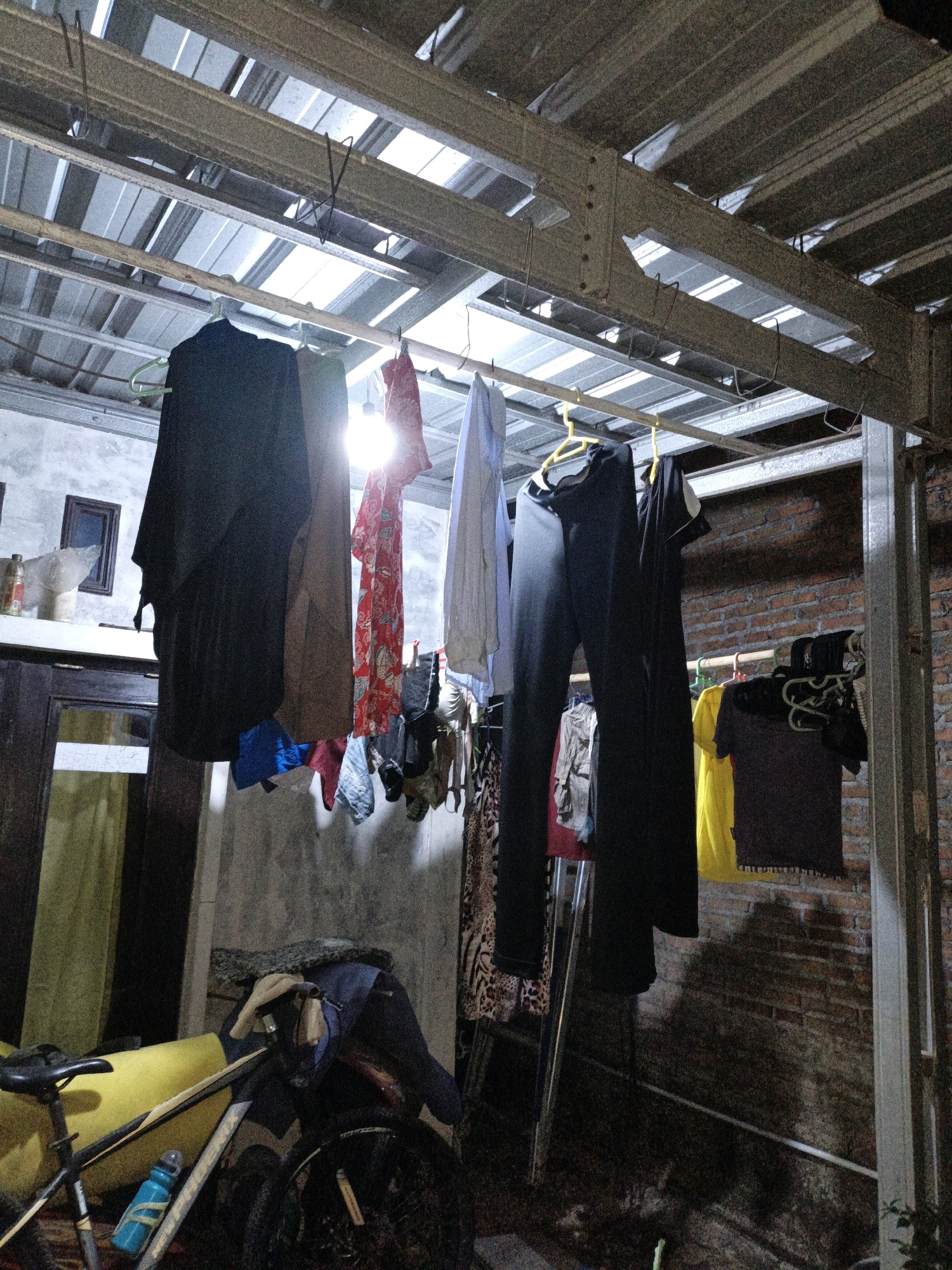 The image depicts a cluttered outdoor area, possibly a backyard or service area, under a metal roof structure. Several items of clothing are hanging on clotheslines, including a black dress or robe, a red and white patterned garment, and various other shirts and pants. Some of these clothes are on hangers, while others are directly on the lines. In the background, there is a brick wall and a window. To the left, a bicycle with a yellow frame and a blue water bottle is visible, along with some other storage items. The lighting appears to be from a single overhead bulb, suggesting it is either evening or an indoor/covered area. The overall scene suggests a utilitarian space used for laundry or storage in what appears to be a residential setting in Cikal, Indonesia. There are no people visible in the image.