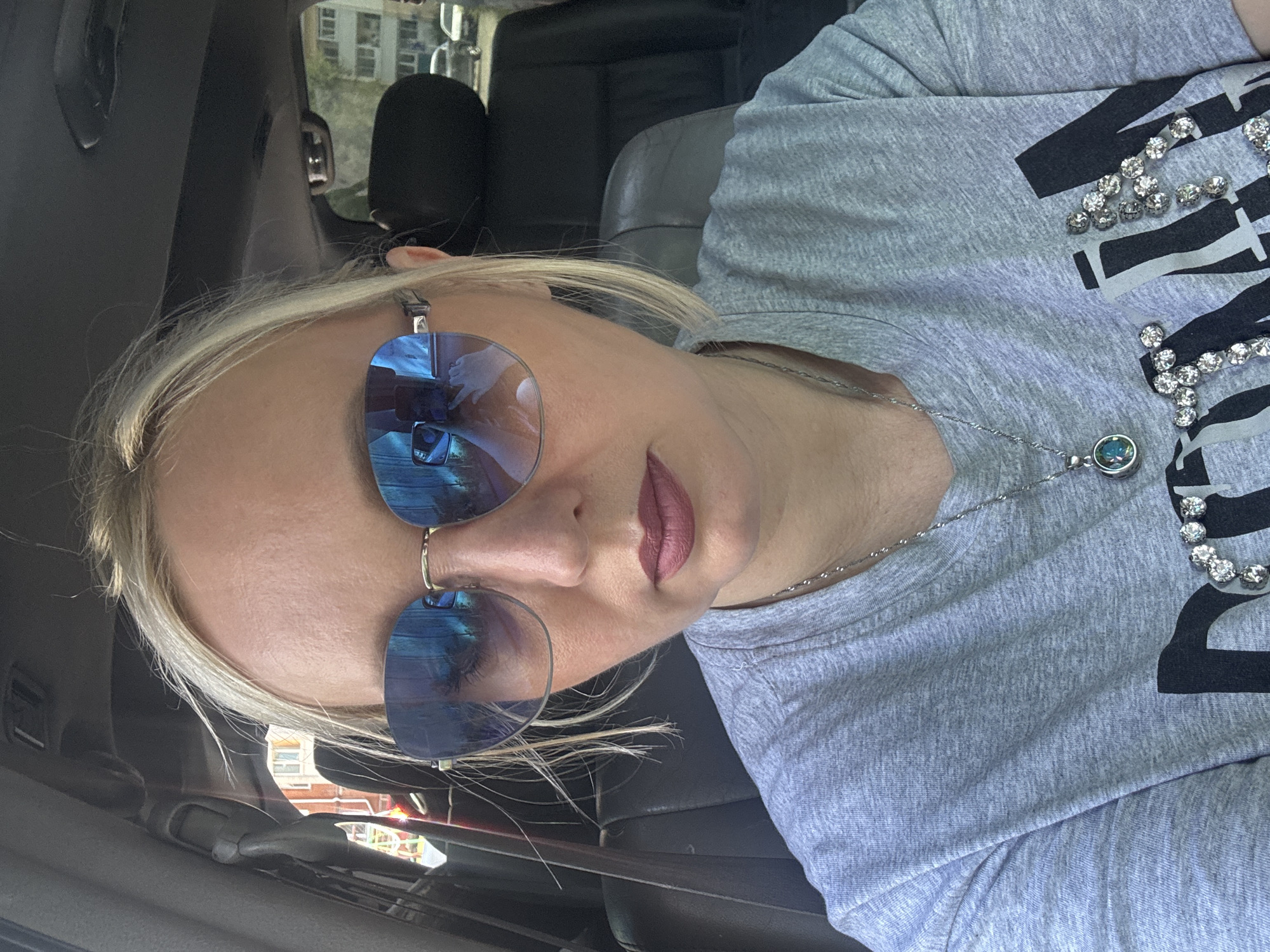 A blonde woman with sunglasses is sitting in a car, looking at the camera. The woman is wearing a gray t-shirt with a necklace. The car appears to be parked in a city, with buildings visible in the background. The context of Komsomolsk-on-Amur, Russia is not evident from the image itself.