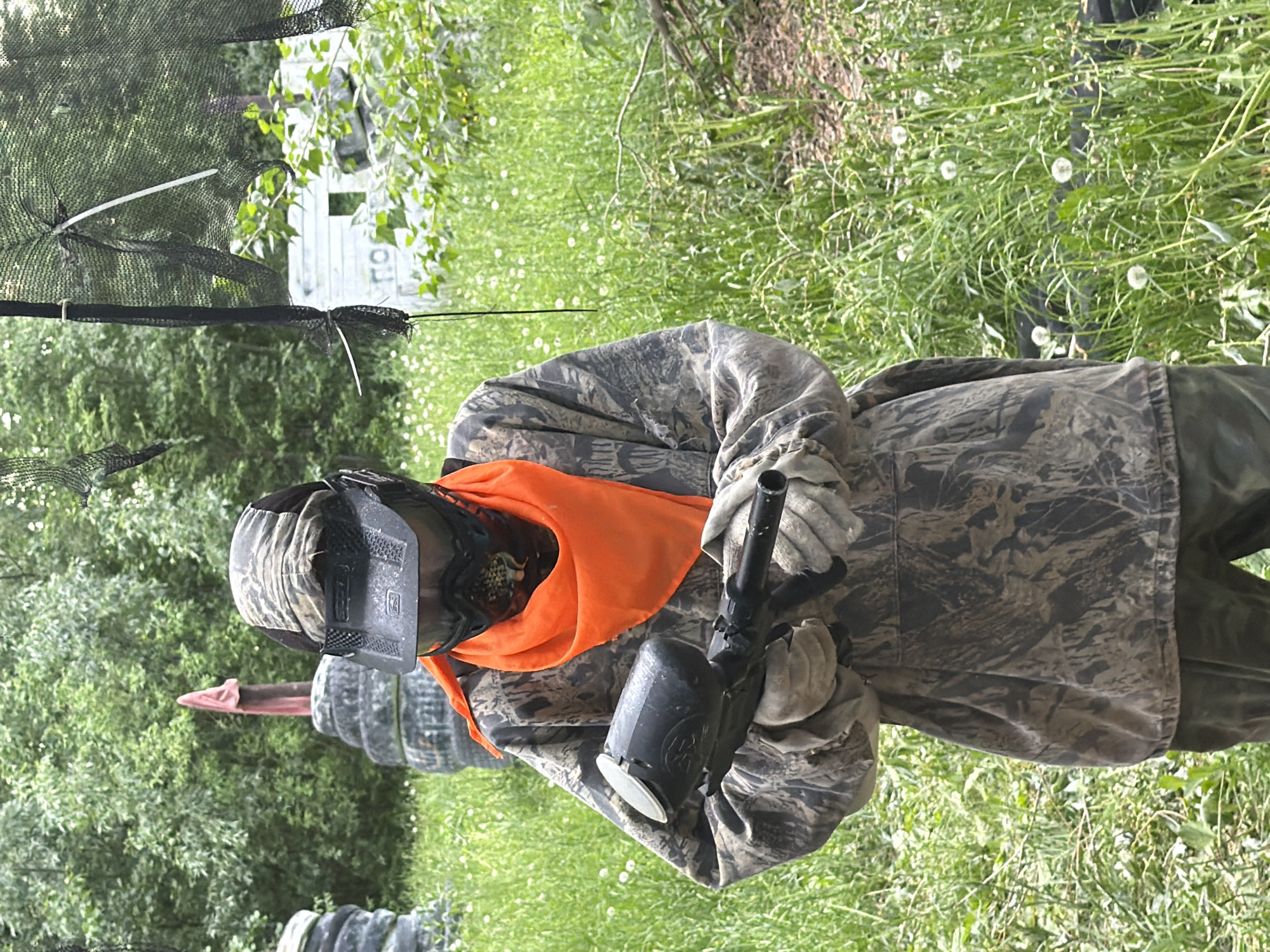 A person in camouflage clothing and a paintball mask stands outdoors, holding a black paintball gun. They also wear an orange bib. The location appears to be a paintball field with green grass, trees, netting, and inflatable bunkers. The specific city is not determinable from this image.