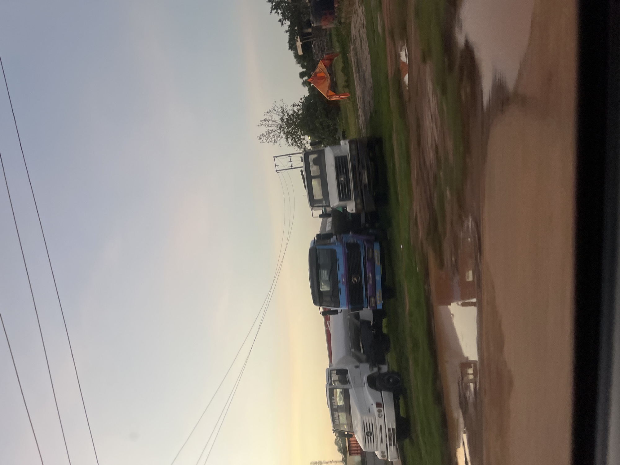 The image depicts an outdoor scene in Galadimawa Village, Nigeria, during what appears to be late afternoon or dusk, characterized by a light blue to grayish-white sky with an orange/yellowish tint near the horizon. Multiple dark horizontal power lines traverse the upper portion of the sky.
In the midground, three large tank trucks are parked on uneven ground consisting of reddish-brown dirt/mud and patches of green grass.
The leftmost truck is white, identified as a Mercedes-Benz, with a white cylindrical tank mounted on its chassis. The text "NAIMA HAKA" is discernible on its sun visor area.
Adjacent to it, a second truck features a blue and purple painted cab with a dark tank. This truck also appears to be a Mercedes-Benz.
The third truck, positioned further to the right, has a white cab and a dark tank; it also appears to be a Mercedes-Benz.
Behind the trucks, a dark metal utility pole structure is visible, supporting additional power lines. Sparse green vegetation, including trees and bushes, is present in the background. An orange or red tarp-like structure is visible on the far right side of the image, behind the rightmost truck. Faint outlines of other distant structures or debris are present further back.
The foreground is dominated by reddish-brown muddy ground with several large puddles. These puddles reflect portions of the sky and the lower sections of the parked trucks, showing distorted reflections of their chassis and tanks. A dark, blurred horizontal band, consistent with the interior of a vehicle window frame, occupies the very bottom of the image. No individuals are visible within the frame, and no specific actions or interactions are occurring.
