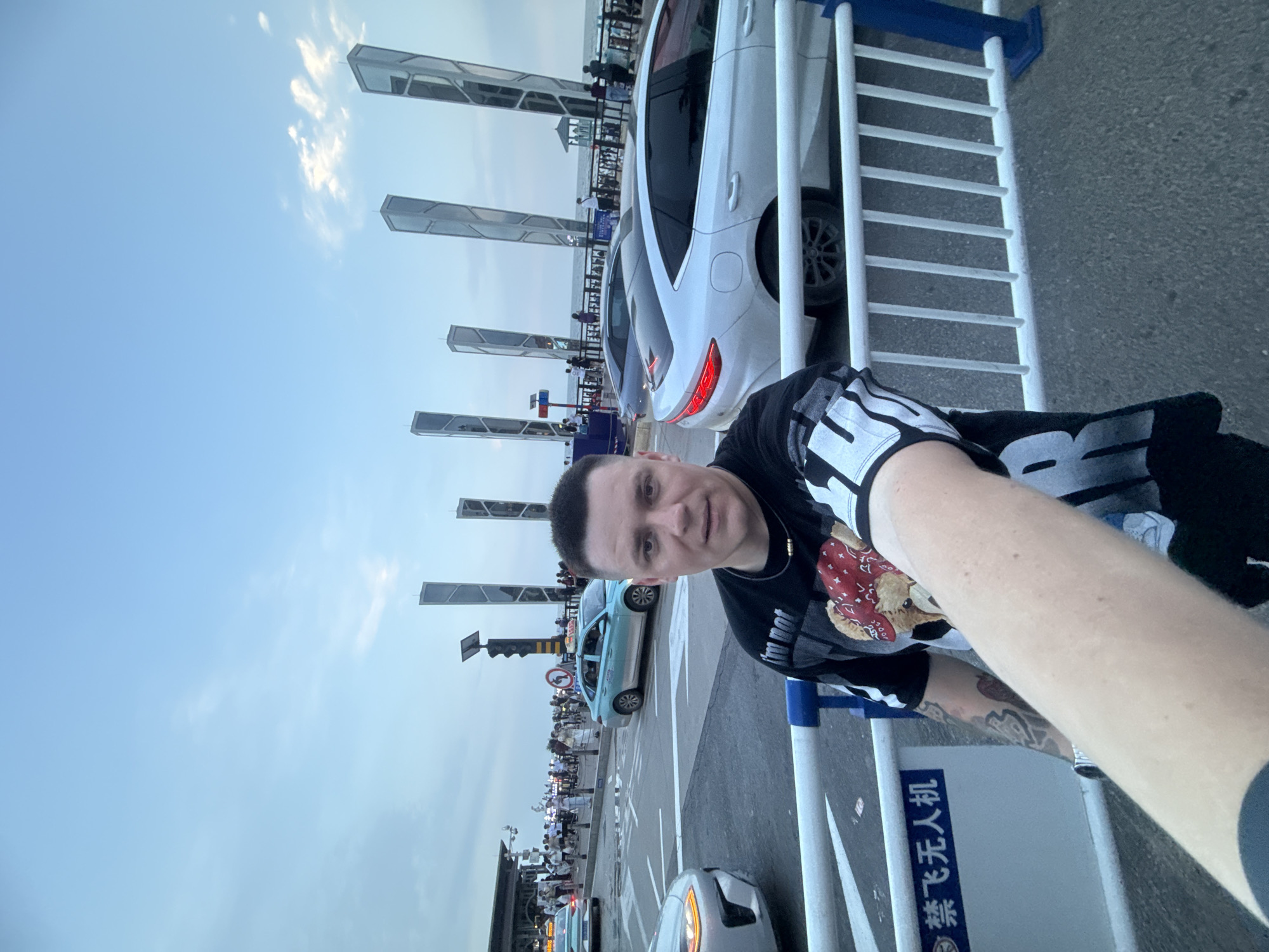 A person is taking a selfie on a street in Qinhuangdao, China, near the beach. They are wearing a black t-shirt with a graphic print. In the background, cars are driving on the road, and there are several tall, modern sculptures. Pedestrians are visible on the sidewalk. The sky is overcast, and the overall lighting suggests it is late afternoon or early evening. A sign with Chinese characters is visible on a white barrier.