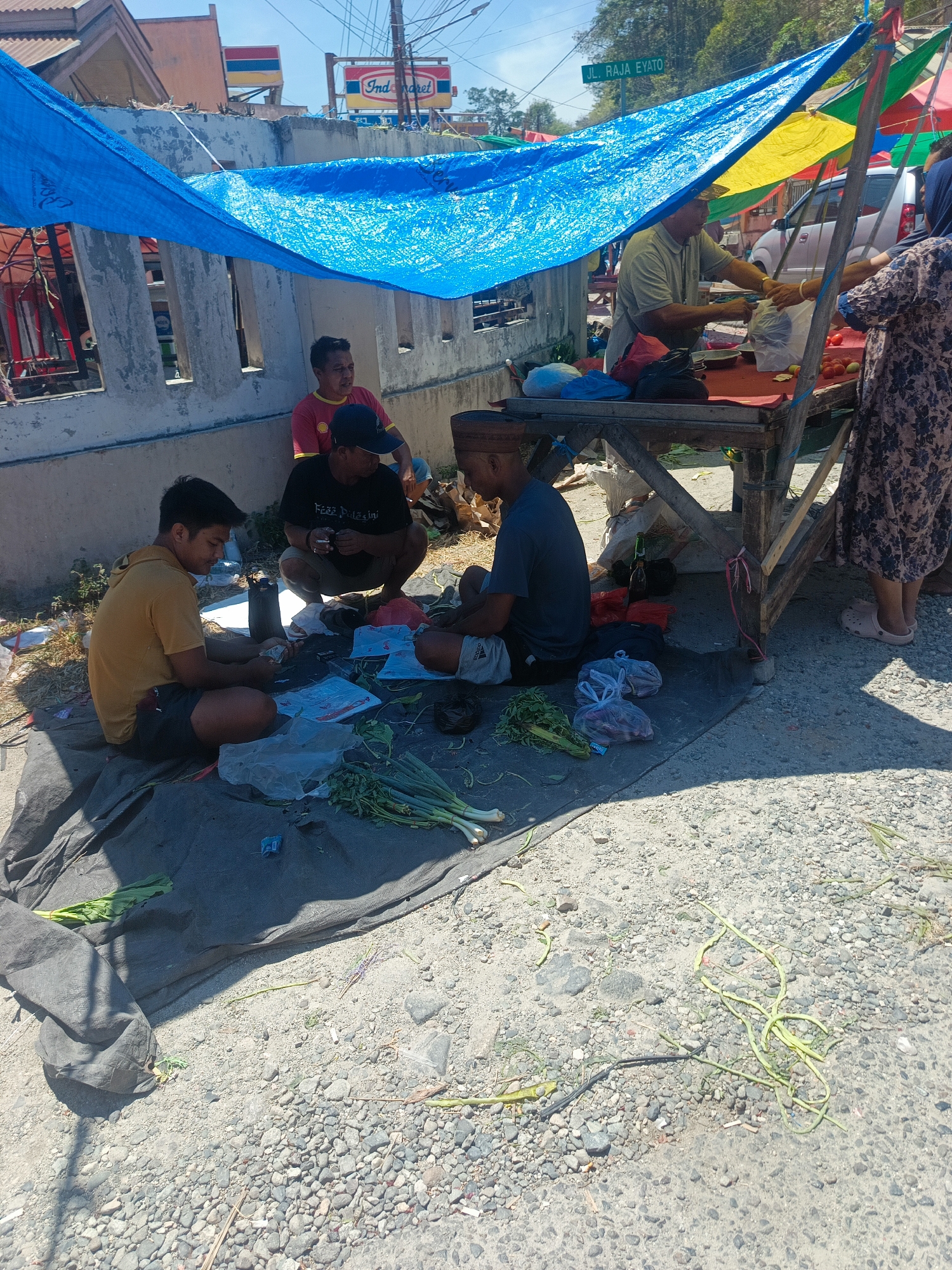 A street-level view captures a modest outdoor market stall under a blue tarp, likely situated in Gorontalo, Indonesia, as evidenced by the street sign "JL. RAJA EYATO". The stall is rustic, constructed from wooden planks. Three men are present: one in a yellow shirt and dark shorts sits on a grey tarp on the ground, engaged with something in his hands; another man in a red shirt and dark shorts, and a third man in a black t-shirt and khaki shorts also sit on the ground, facing the stall. A woman in a patterned dress and headscarf stands at the stall, interacting with a fourth individual, an older man in a light grey shirt and a hat, who appears to be a vendor. He is reaching out, possibly handing over items or making a transaction. The ground is a mix of gravel and dirt. In the background, a wall and a building with an "Indomaret" sign are visible, suggesting a commercial area. The scene is bathed in bright sunlight, casting shadows. Various items, including produce like green onions and tomatoes, are displayed on and around the stall, along with plastic bags and containers.