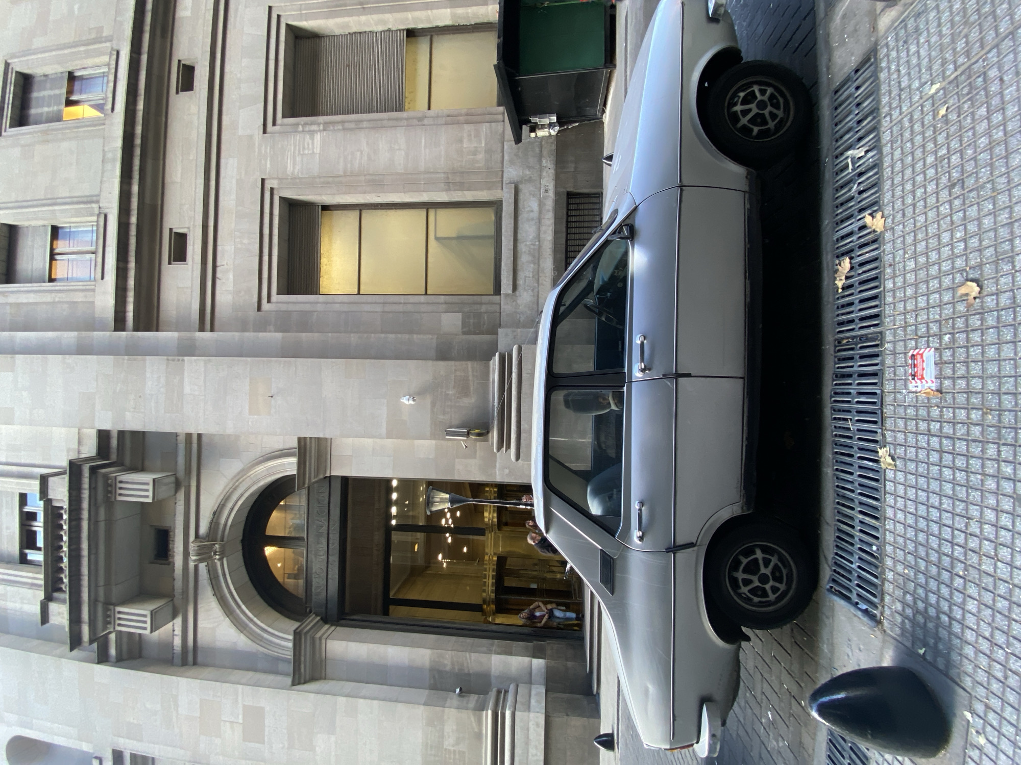 Here is a description of the image:
Exterior shot. A light silver-colored 1970s-era sedan is parked directly in front of the main entrance of the Banco de la Nación Argentina in Buenos Aires, Argentina. The neoclassical architectural features of the bank building are evident, including a large arched entranceway, several upper-level windows, and the building's stonework. People are entering and exiting the building. The street is paved and contains a metal grated drain. The time of day appears to be daytime, with plenty of natural light.