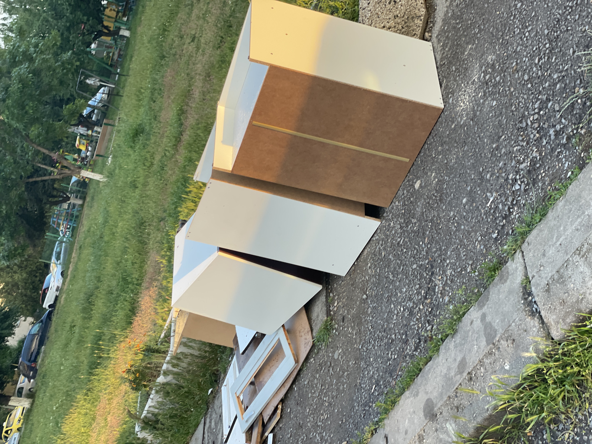 The image shows a discarded pile of disassembled furniture, likely cabinets or drawers, on a paved surface next to a grassy area. The furniture is made of wood and MDF, with a white finish on the front panels. The scene is outdoors, potentially on a curb or near a sidewalk. Based on the architecture and vegetation visible in the background, the location could be in a residential area in Sofia, Bulgaria. There are no people visible in the image. The objects include wooden furniture components, concrete pavement, grass, and trees. The environment suggests an urban or suburban setting with green spaces.