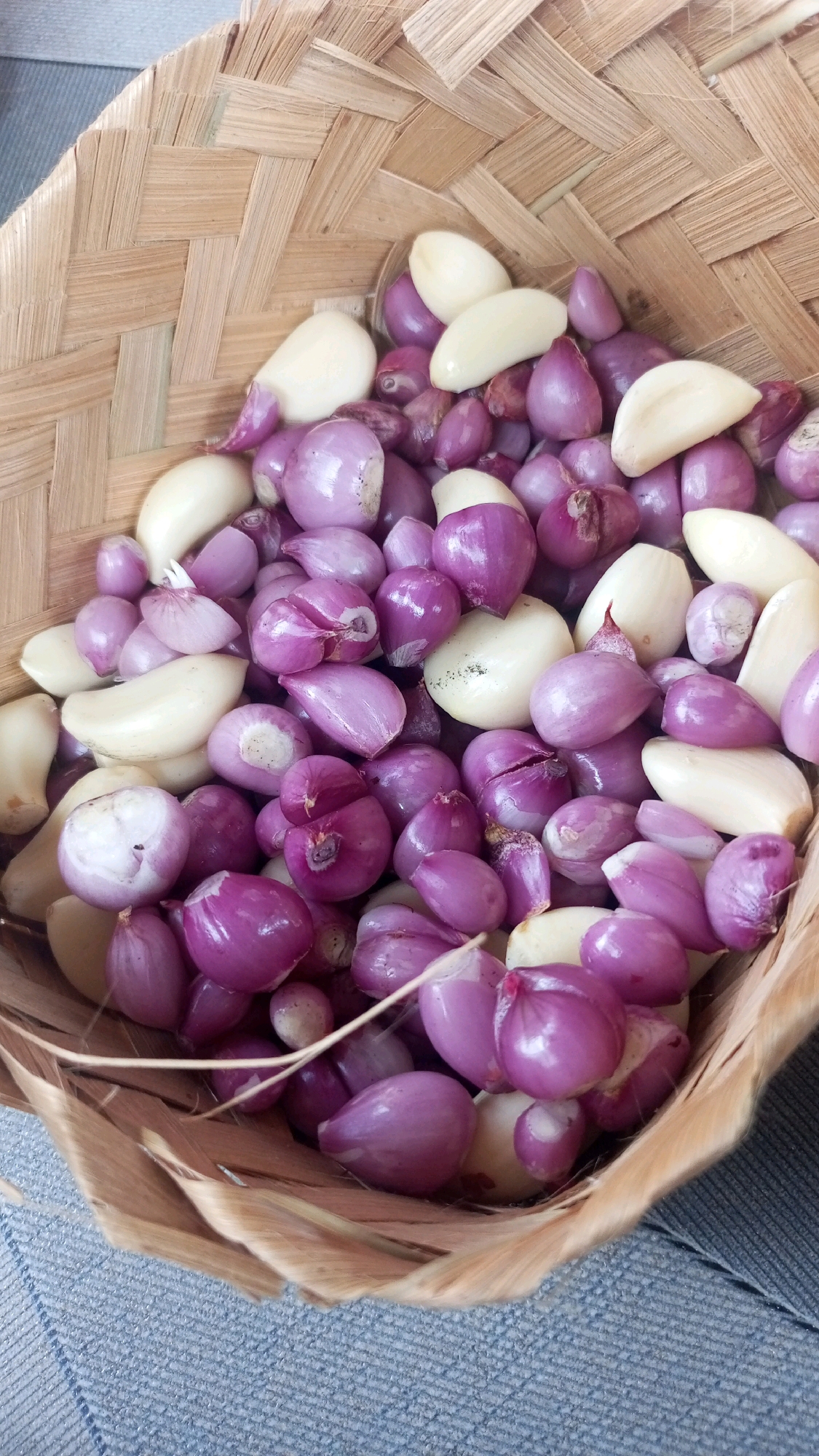 A close-up, overhead shot shows a woven basket filled with shallots and peeled garlic cloves. The shallots are small and predominantly purple, with some having a lighter hue. The garlic cloves are white and appear to be of various sizes. The basket is made of light brown woven material, with the texture of reeds or bamboo visible. The basket is placed on a textured surface that appears to be blue denim. The image was taken in the city of Mangliawan, Indonesia.