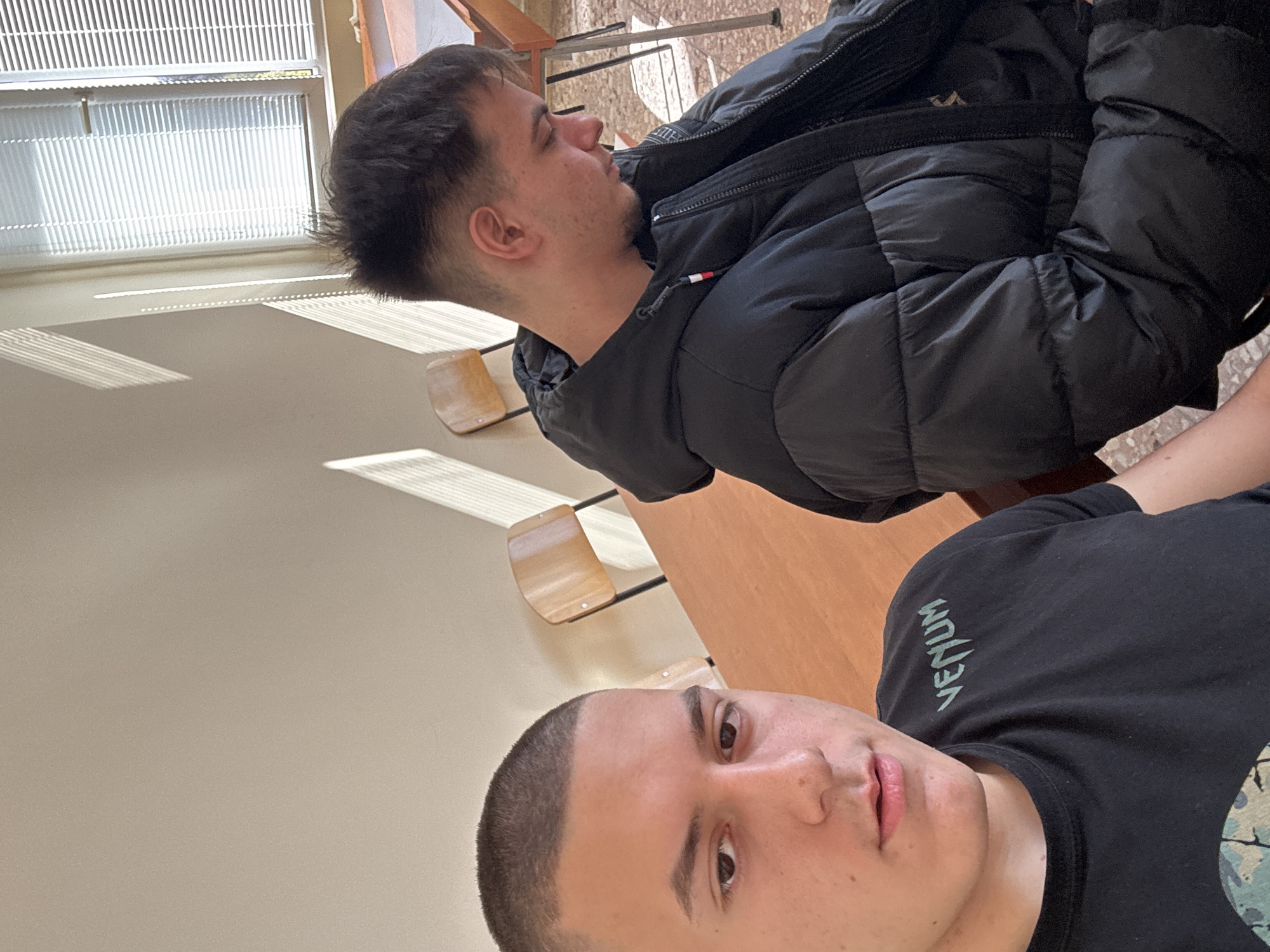 The image depicts two young men indoors. One is facing the camera, wearing a black t-shirt with the word "VENUM" printed vertically. The other is to his left, facing away from the camera and wearing a black puffer jacket. They are seated at wooden desks, which are common in classrooms. Light enters the room through a window with blinds. The location is likely in Plovdiv, Bulgaria based on the prompt. The setting appears to be an educational institution or classroom.