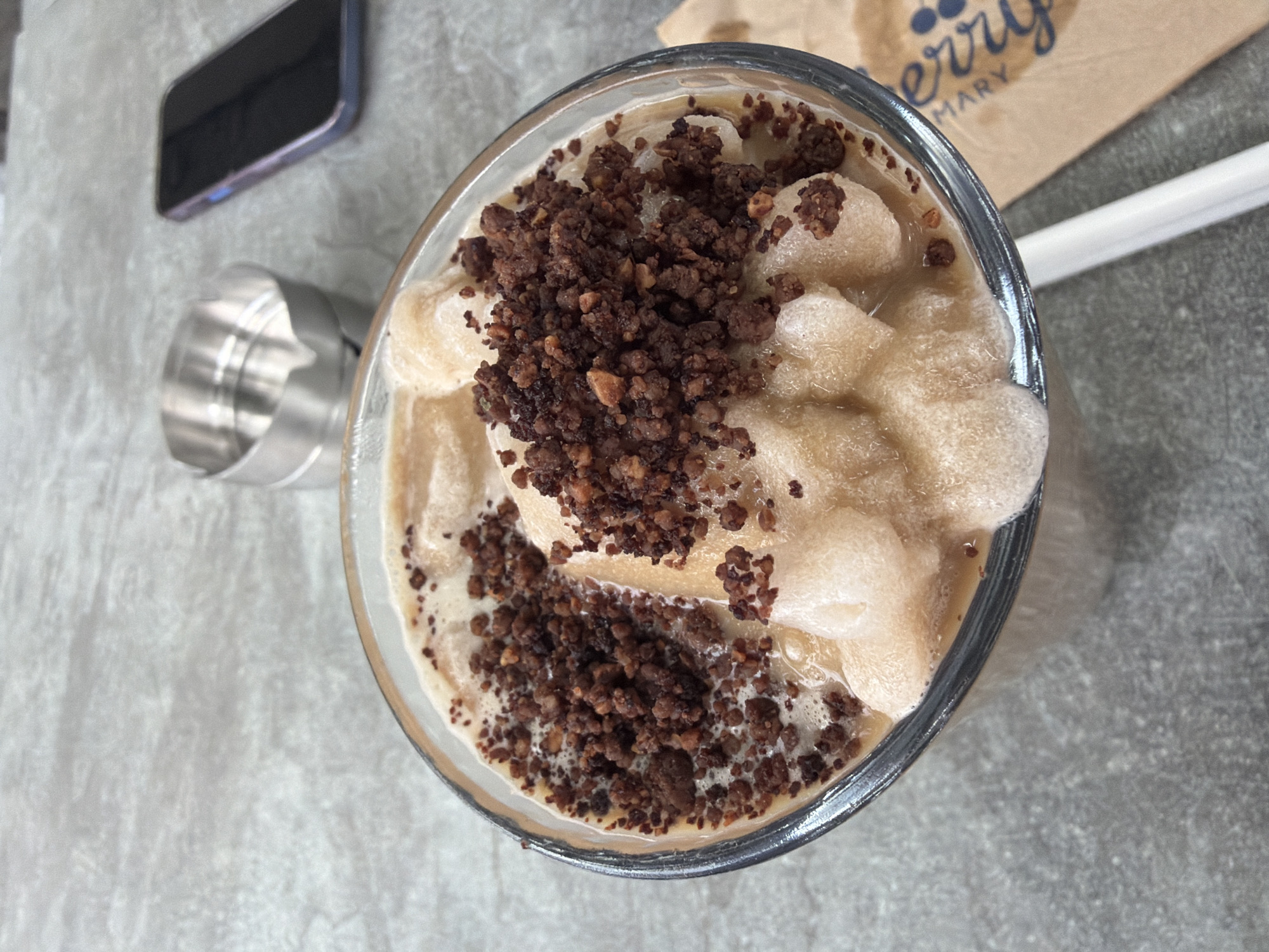 A high-angle shot depicts a glass of a blended beverage, possibly a coffee or smoothie, topped with granular dark brown sprinkles. The drink sits on a gray, textured surface alongside a stainless steel measuring cup, a black smartphone, a brown paper napkin with partial lettering, and a white straw. The scene appears to be staged on a table in a cafe or similar establishment. The location cannot be determined as Varna, Bulgaria with certainty from the available visual information.