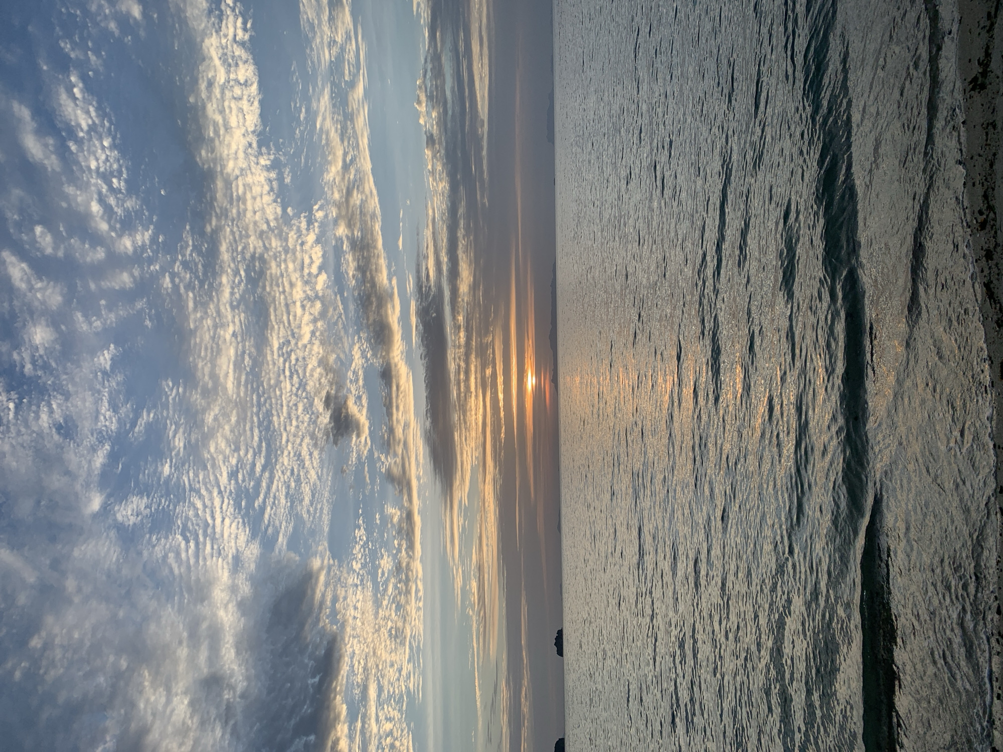 A serene sunset over the ocean is captured in this image, likely taken from a beach in Ko Samui District, Thailand. The sky is a canvas of soft blues and greys, with a band of delicate, wispy clouds stretching across the horizon, illuminated by the warm glow of the setting sun. The sun itself is a small, bright orb peeking just above the distant water, casting a shimmering golden pathway across the gently rippling surface. In the far distance, several small, silhouetted islands break the monotony of the ocean. The foreground shows a narrow strip of sandy shore, where gentle waves lap against the beach. The overall atmosphere is one of tranquility and natural beauty, with no people or specific man-made structures immediately visible.
