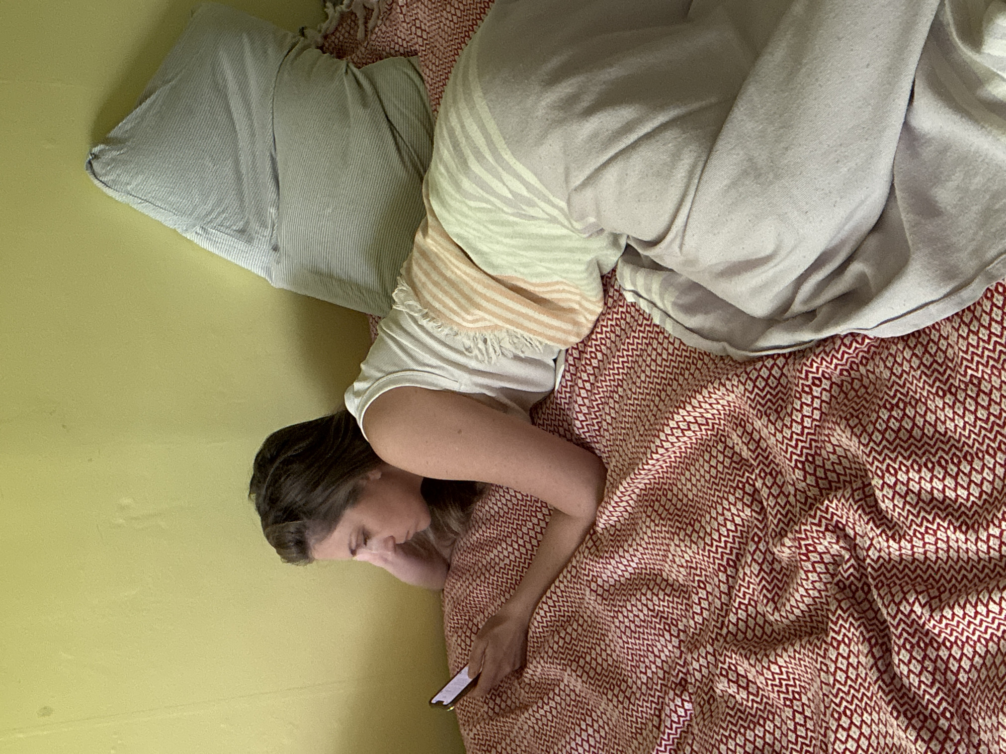 A woman is lying on a bed, looking at a cell phone in her hand. She is on her side, with her head near a pillow and part of her body covered by a blanket. The bed has a red and white patterned sheet or blanket. The wall beside the bed is a pale yellow color. The location is unknown and cannot be determined from the image.