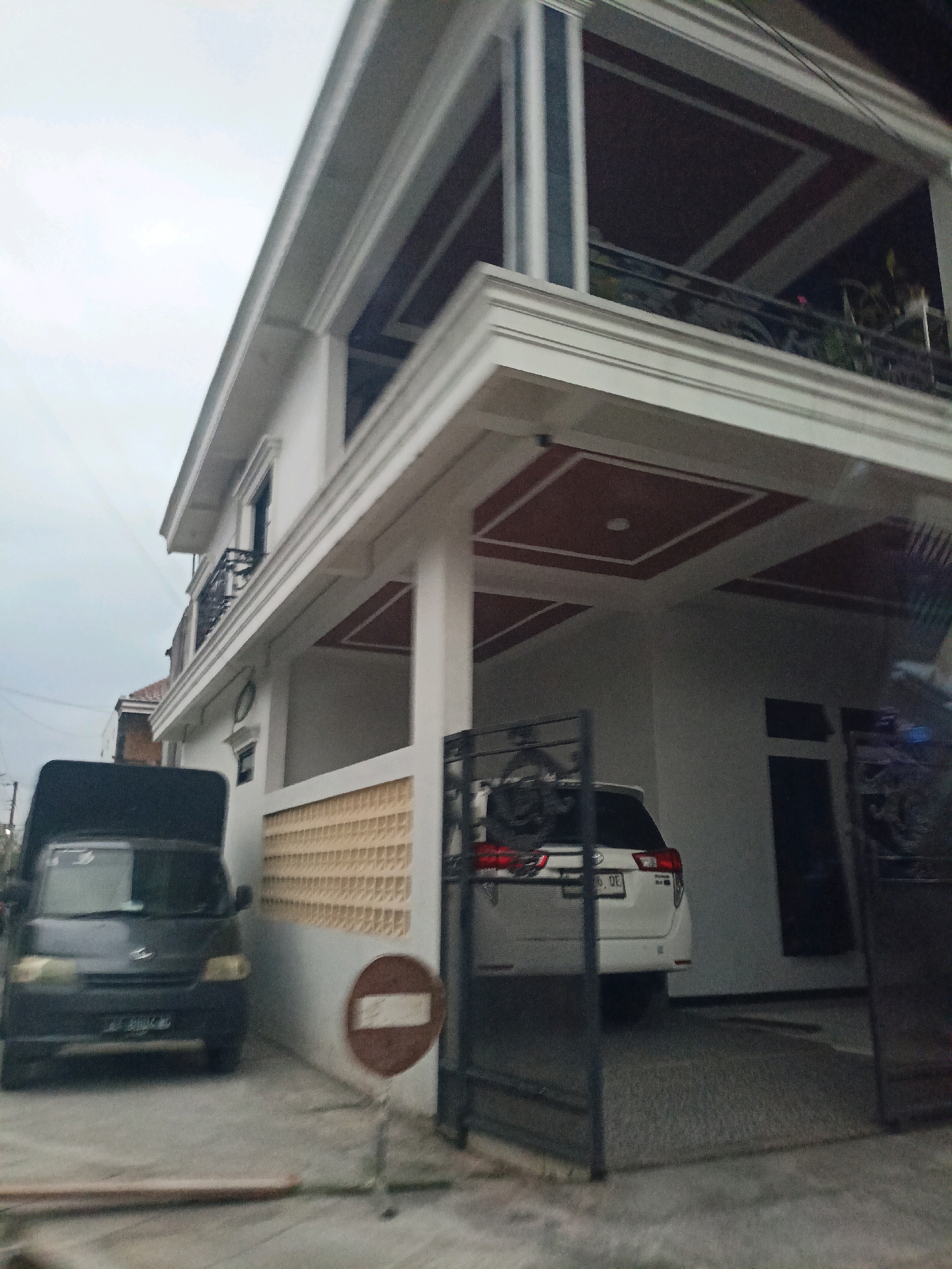 A wide shot of a modern, two-story house in the city of Payaman, Indonesia, on a cloudy day. The house is white with dark red accents on the ceilings of its covered porches and balconies. A black pickup truck is parked on the left side of the image, facing the house. To the right of the truck, a white SUV is parked in the driveway. A metal gate is visible in front of the driveway. A round, brown and white traffic sign is placed on a pole in front of the house. The overall scene is well-lit, suggesting it is daytime.