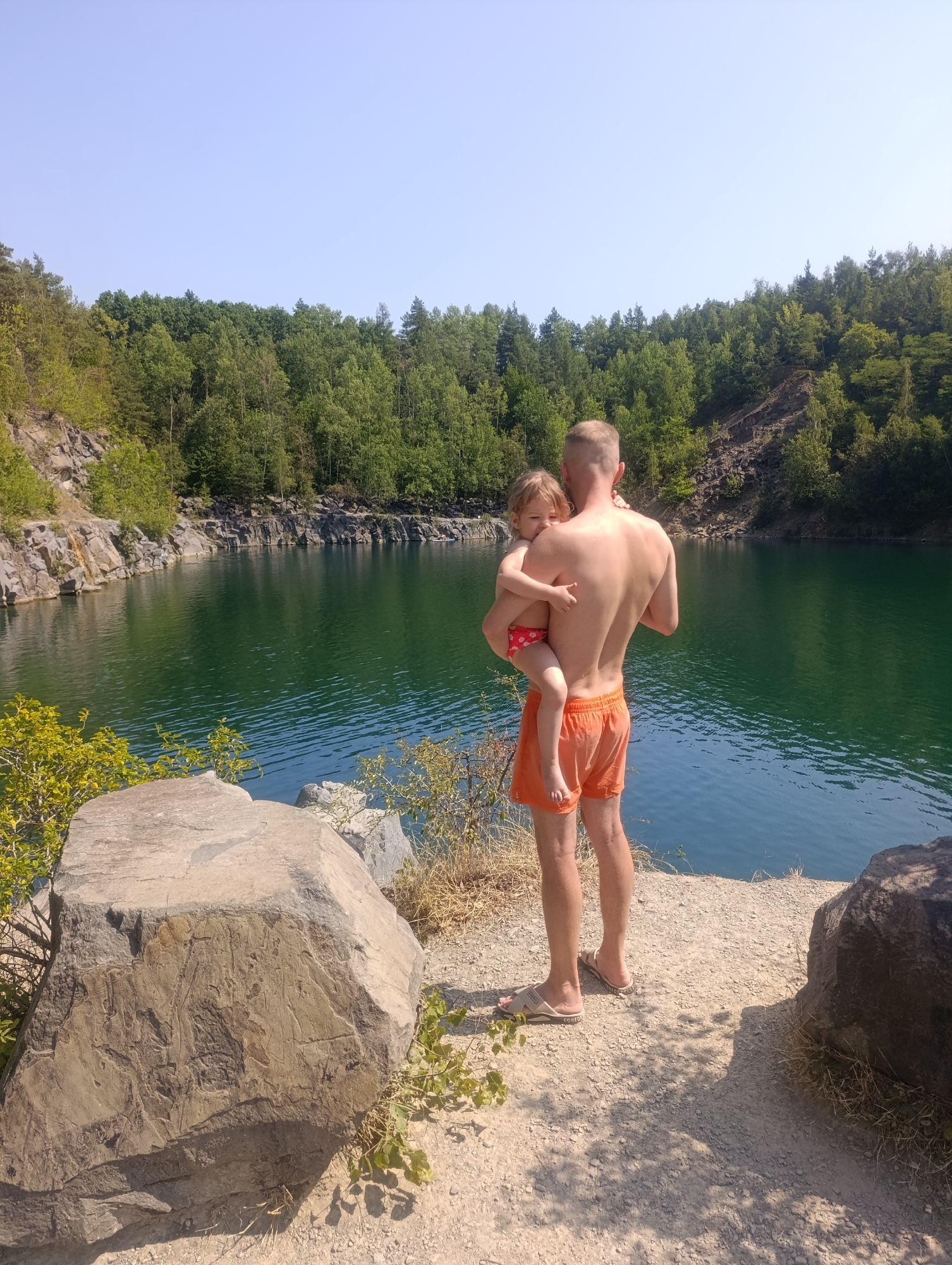 A father stands on a rocky shore, holding his young daughter. He is wearing orange swim trunks, and his daughter is in a red and white patterned swimsuit. The lake before them is a deep emerald green, reflecting the lush green trees that line the opposite shore. Rocky cliffs rise behind the trees, suggesting a quarry or a natural rock formation. The sun is high in the sky, casting sharp shadows and indicating it is midday. The weather appears to be warm and clear, perfect for a swim. The scene evokes a sense of peace and family enjoyment in a natural setting. There is no visible text in the image.