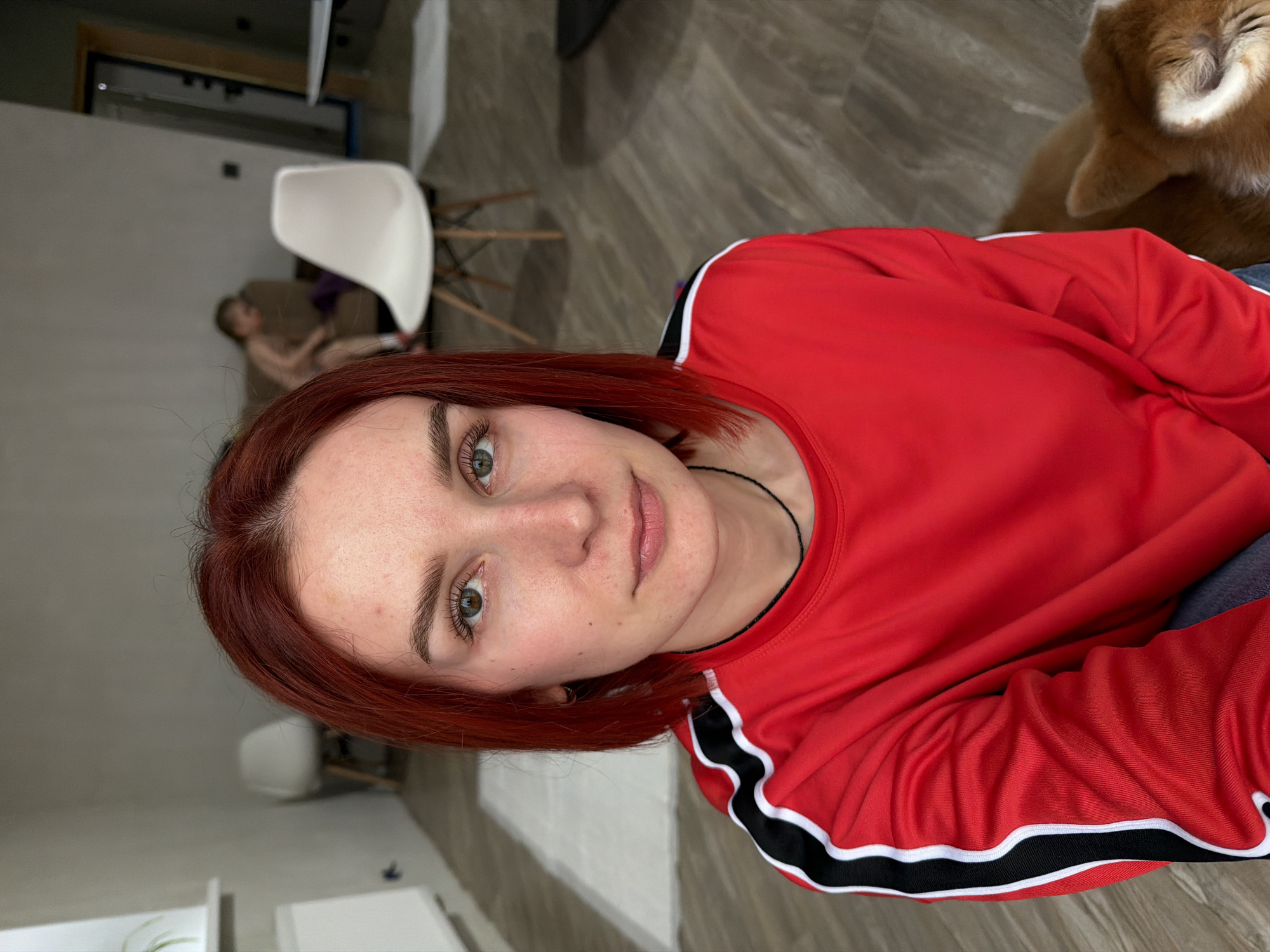 A red-haired woman in a red sports shirt with black and white stripes is taking a selfie. Her hair is shoulder-length and red, and she has green eyes. A dog is in the upper-right corner, and a person is blurred in the background near a white chair. The location appears to be indoors, possibly a home, in what could be Chaikovsky, Russia.