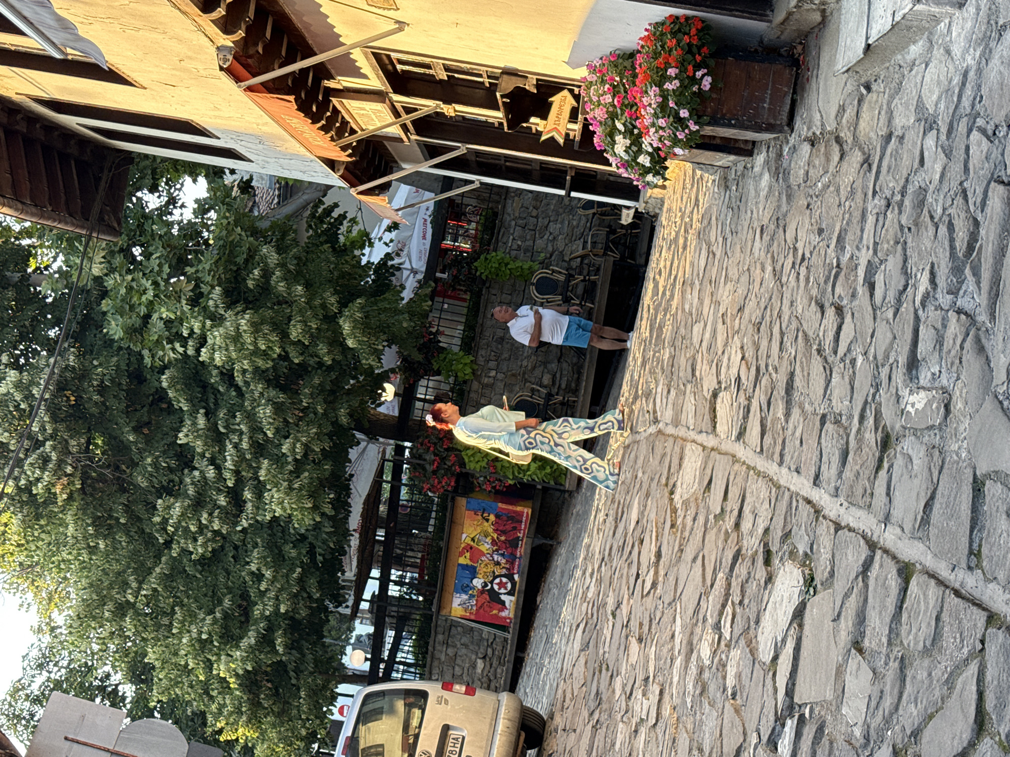 The image depicts a cobblestone street in Veliko Tarnovo, Bulgaria, likely in the historic old town given the architectural style and the presence of a colorful mural. In the foreground, a woman in a light-colored, patterned outfit walks away from the camera, her blonde hair tied back. To her right, a man in a white polo shirt and blue shorts stands, holding a phone. Behind them, a silver van is parked on the left. The street is lined with buildings featuring traditional architecture with wooden beams and textured walls. Colorful flowers in wooden planters add a vibrant touch to the scene. The overall atmosphere suggests a warm, sunny day, possibly late afternoon, with long shadows cast by the buildings and trees. The mural behind the woman appears to be political or historical in theme, with figures and symbols.