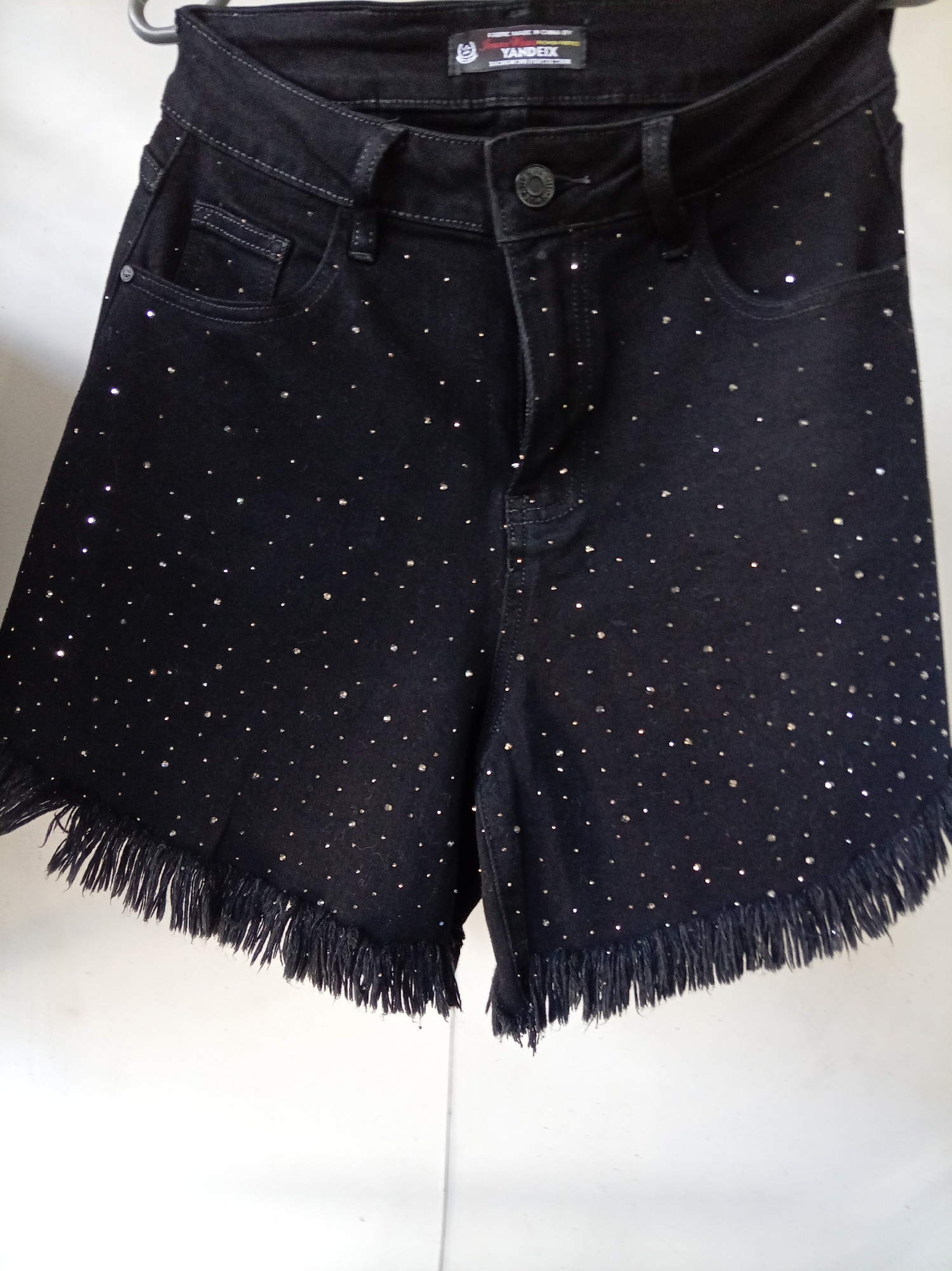 A pair of black denim shorts with a frayed hem and scattered rhinestones is displayed on a hanger against a white background. The shorts have a zip fly and button closure. A brand tag reading "YANDEIX" is visible at the waistband. The lighting suggests an indoor setting, potentially a retail display or a home closet. The image shows no signs of the city of Donetsk, Ukraine, or any specific environmental context outside of the object itself.