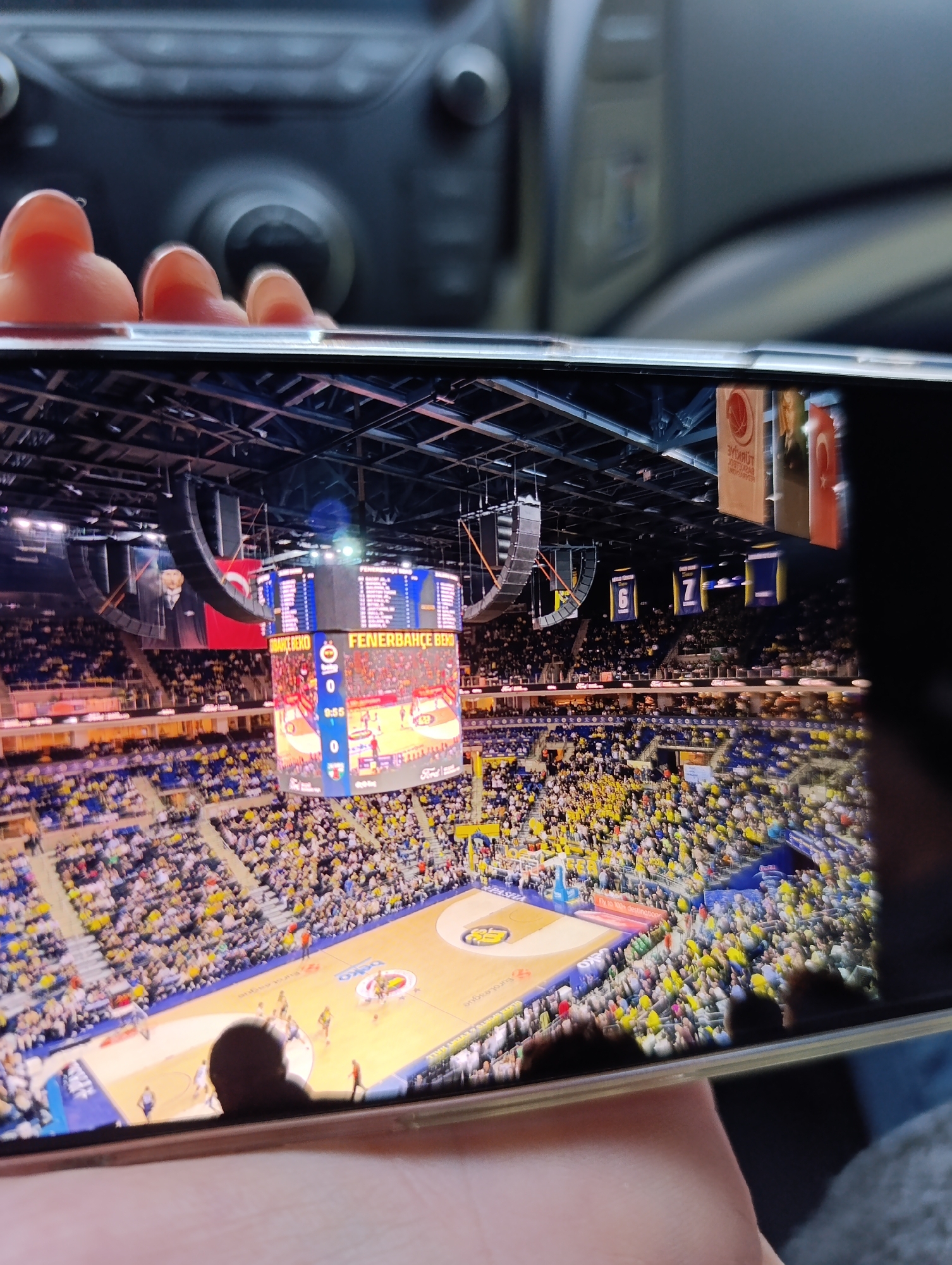 The image shows a smartphone screen displaying a basketball game in a large arena with many spectators. The basketball court is visible with players in action. A large scoreboard with the name "FENERBAHCE BEKO" is hanging above the court. Banners and flags are visible in the background. The phone is being held by a person, and part of the interior of a vehicle is visible in the background behind the phone, including knobs and a surface. There is no evidence to suggest this is in Haskovo, Bulgaria.