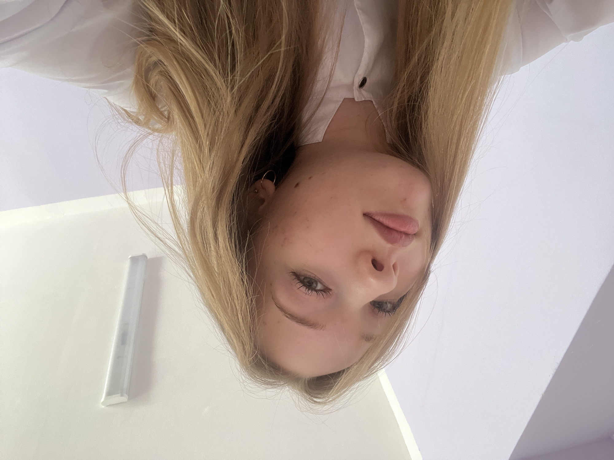 A young woman with blonde hair is looking directly down at the camera. She is wearing a white shirt. Part of a white wall with a white light fixture is visible on the left side of the frame. The background is white or light purple. There is no information in the image to indicate the location is Chaikovsky, Russia.