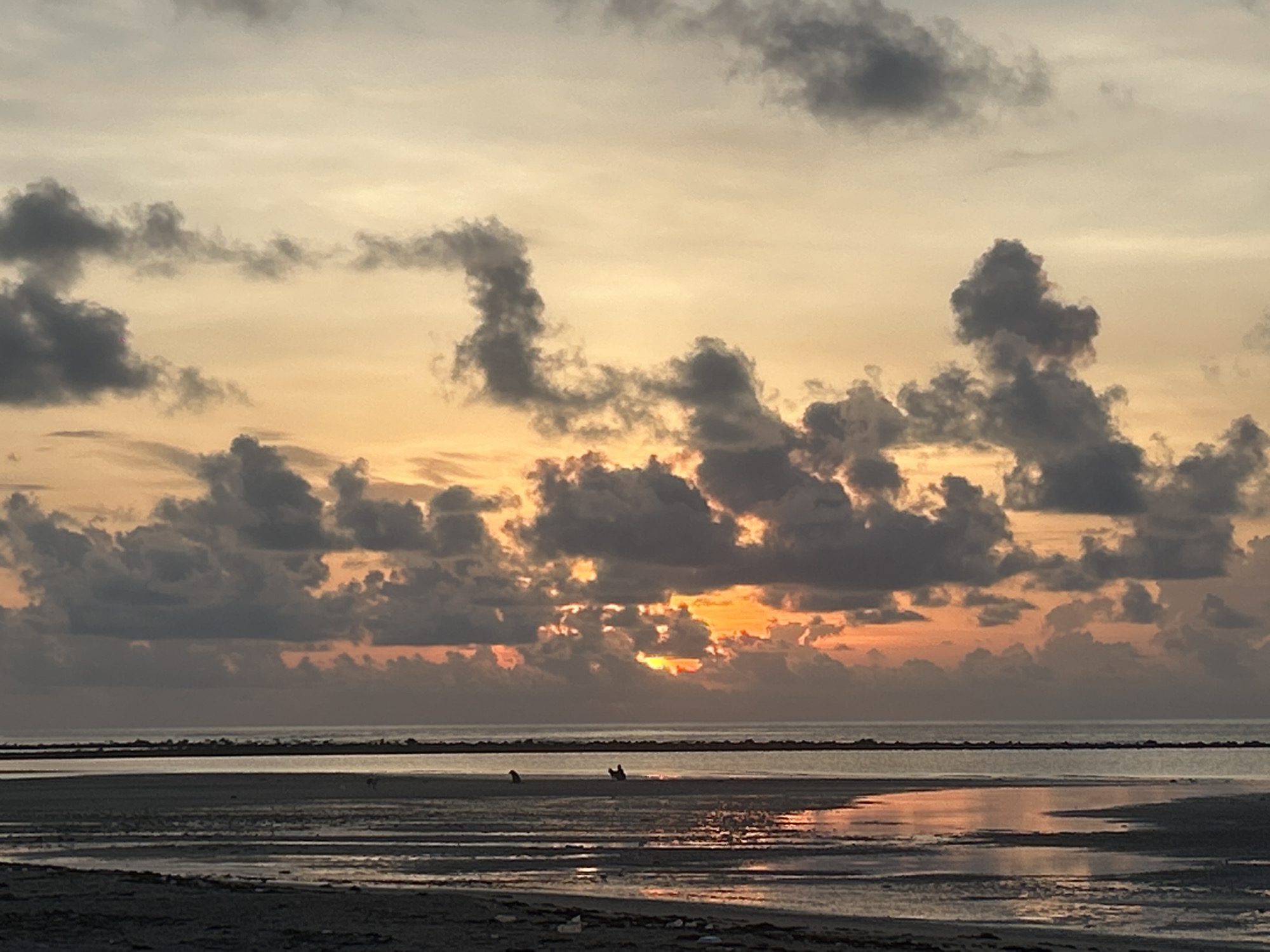 The image depicts a coastal sunset scene. In the foreground, there is a sandy, wet beach area with water reflections. Beyond the beach, a dark line suggests the shoreline or perhaps a distant landmass. Above this line is the sea, its surface somewhat calm. The sky is filled with various cloud formations, some dark and dense, others lighter. The setting sun is partially visible through the clouds, casting an orange glow across the sky. The location is likely Baan Harn, Thailand, given the specified location. There are two figures on the beach. One sitting and one standing. There are two birds on the beach. There is a horizon line between the landmass and the sky.