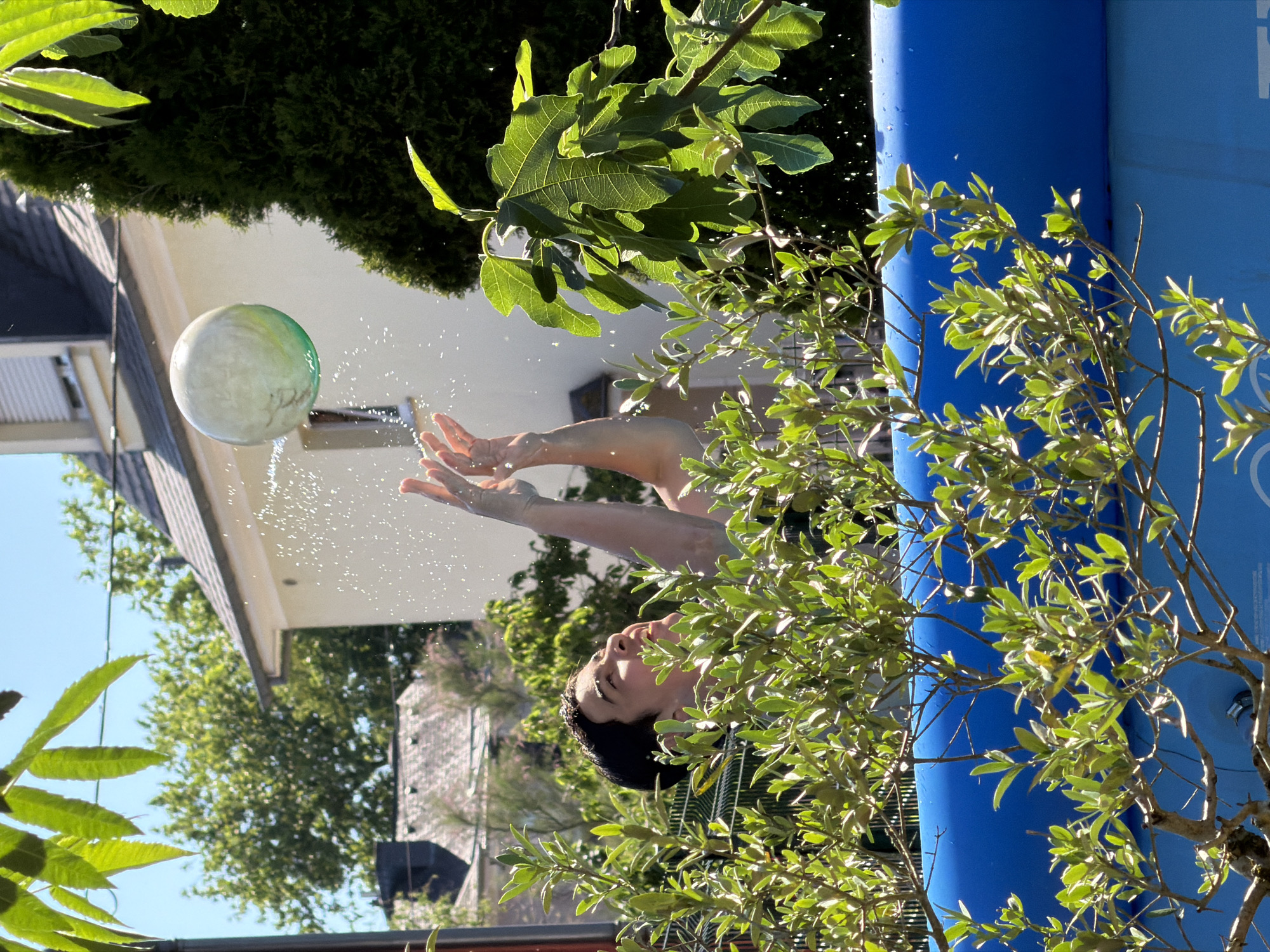 A person is reaching up towards a ball, causing water droplets to splash around it. The scene appears to be outdoors, with a blue inflatable pool in the background, partially obscured by tree branches. The setting includes a residential area, evidenced by buildings and green vegetation. The presence of houses suggests a location in Esch-sur-Alzette, Luxembourg.