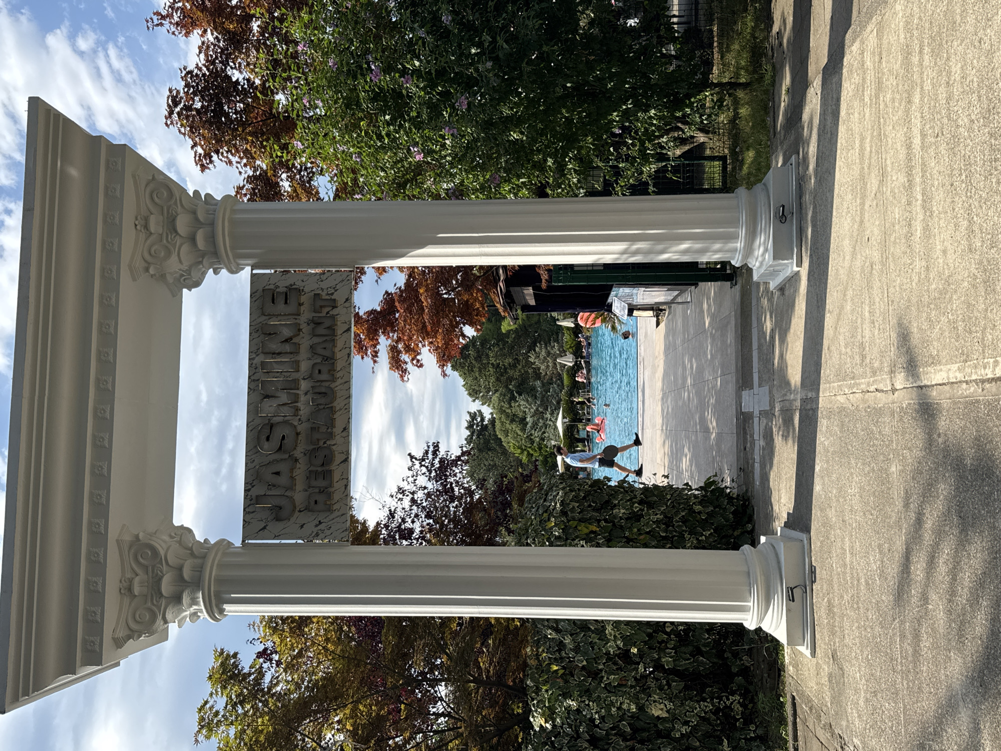 The image shows an entrance to the Jasmine Restaurant in Batumi, Georgia, with a swimming pool area visible behind. The entrance is framed by two white classical-style columns supporting an entablature with decorative molding. A sign hanging between the columns reads "JASMINE RESTAURANT" in raised lettering on a marble-like background.
In the background, a bright blue swimming pool is filled with people, some of whom are in the water, while others are lounging on the side. A large pink inflatable flamingo is floating in the pool. Trees with autumnal foliage in shades of red and orange surround the pool area, contrasting with the lush green foliage of hedges and bushes in the foreground.
A man in a light blue shirt and dark shorts is walking on a paved pathway leading towards the pool. He is carrying a circular object in his right hand. The ground is a mix of concrete slabs and paving stones. Sunlight casts shadows from the columns and trees onto the ground, indicating a bright day. The sky is partly cloudy with a clear blue expanse visible.