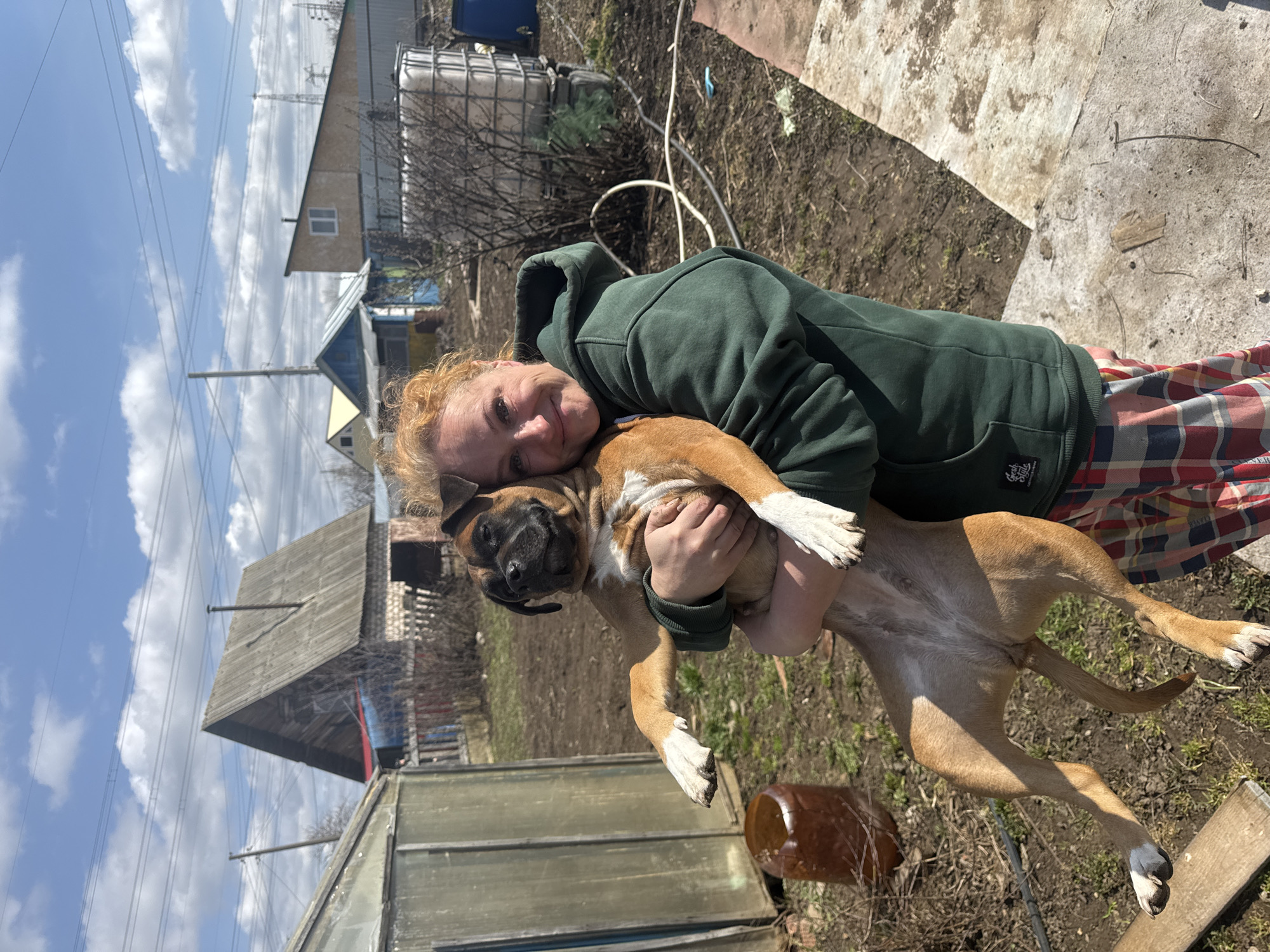 A woman with auburn hair, wearing a green hoodie and plaid pants, is holding a tan boxer dog in her arms. The dog has white markings on its paws and chest. They are outside on a sunny day in a rural or suburban setting. There are sheds and houses in the background, as well as a white water tank. Based on the location information provided, the scene is in Chaikovsky, Russia.