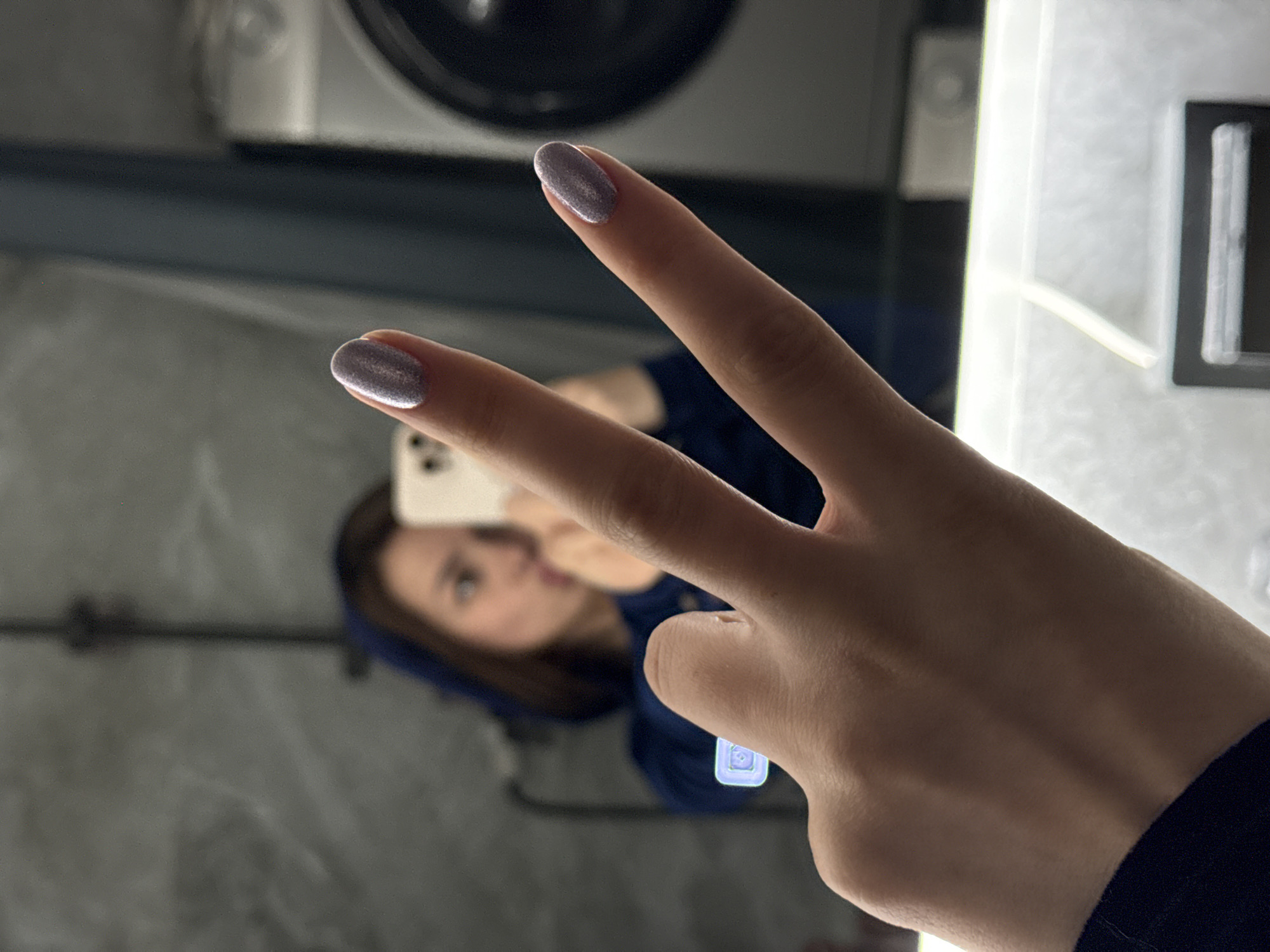A mirrored selfie shows a person holding up two fingers in a peace sign. The hand is in the foreground, with silver nail polish on the fingernails. In the reflection, a woman is visible holding a smartphone. The background includes what appears to be a reflective surface, possibly a mirror or window, showing elements of an interior space. The location, Лобаново, Russia, cannot be verified from the image.